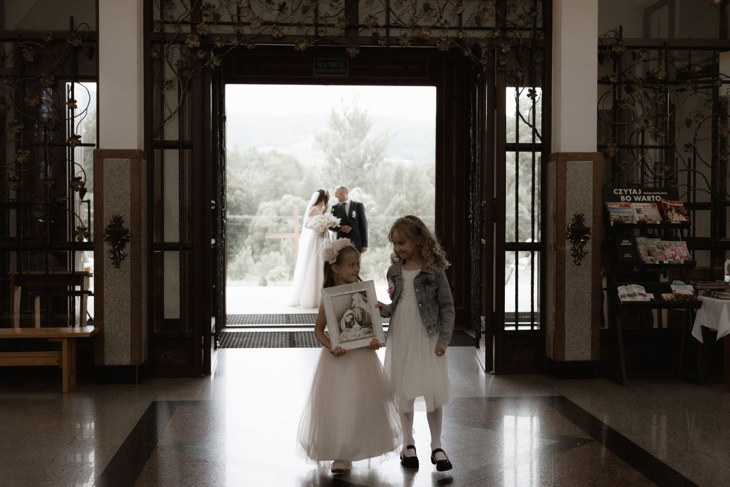 Two young children in formal attire stand silhouetted in the doorway of an elegant venue with ornate details.