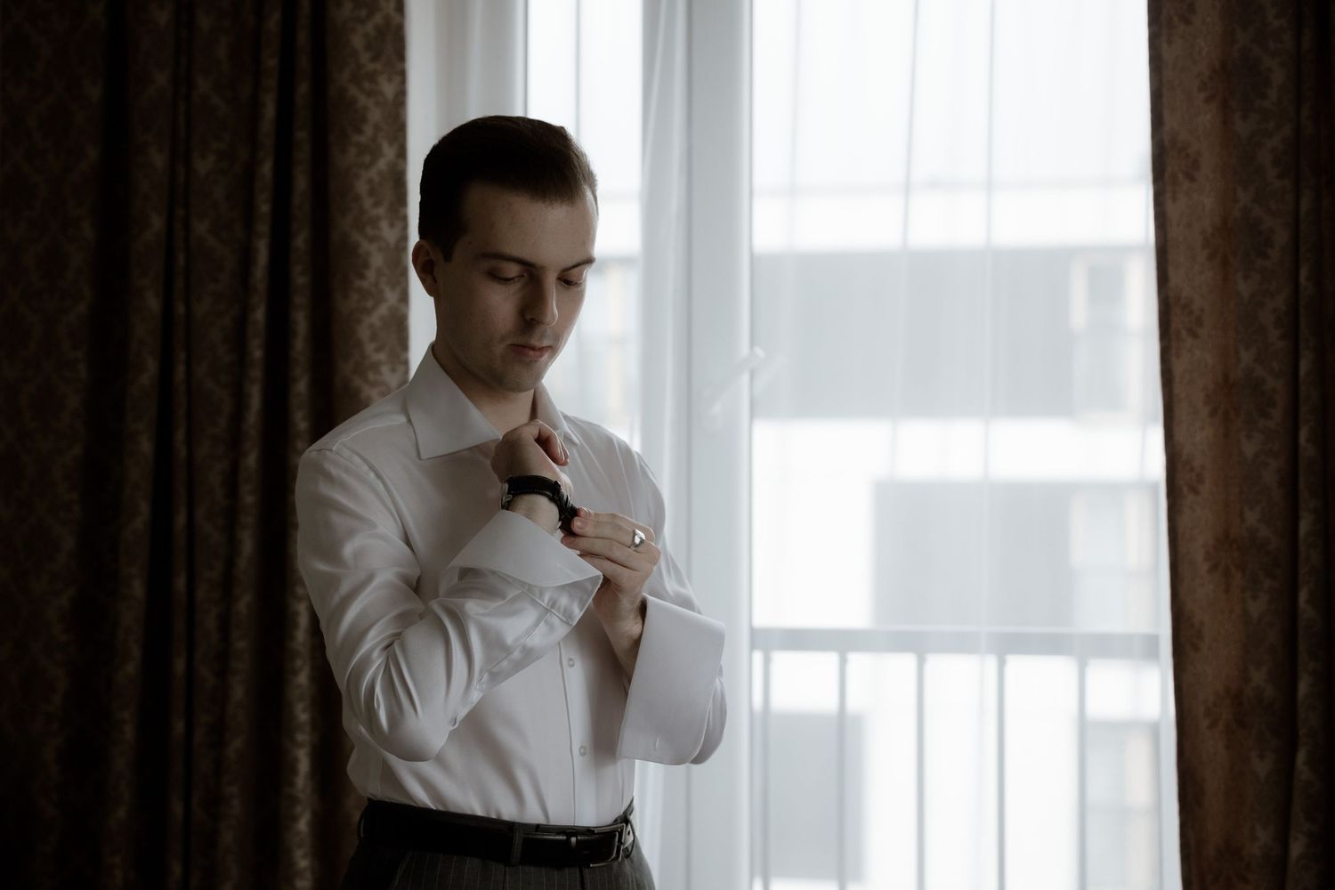 A person in a white dress shirt adjusts their sleeve cuff while standing by a window with dark curtains.
