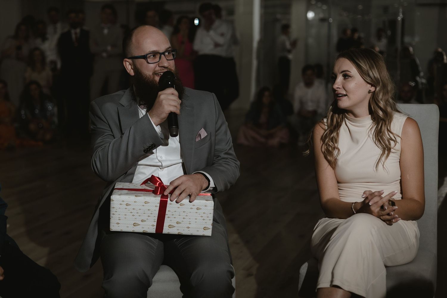 A wedding guest with a beard holds a wrapped gift while sitting next to a person in a white dress at an evening reception.