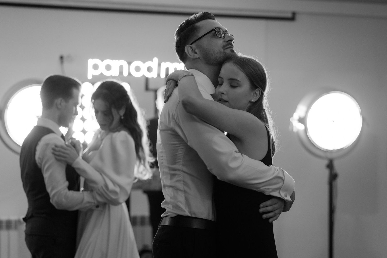 Couples slow dance together in black and white at an indoor event with illuminated circular lights in the background.