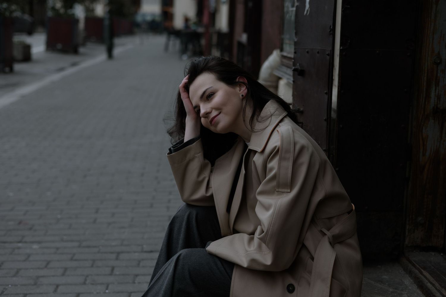 Person in beige coat sitting thoughtfully against urban backdrop.