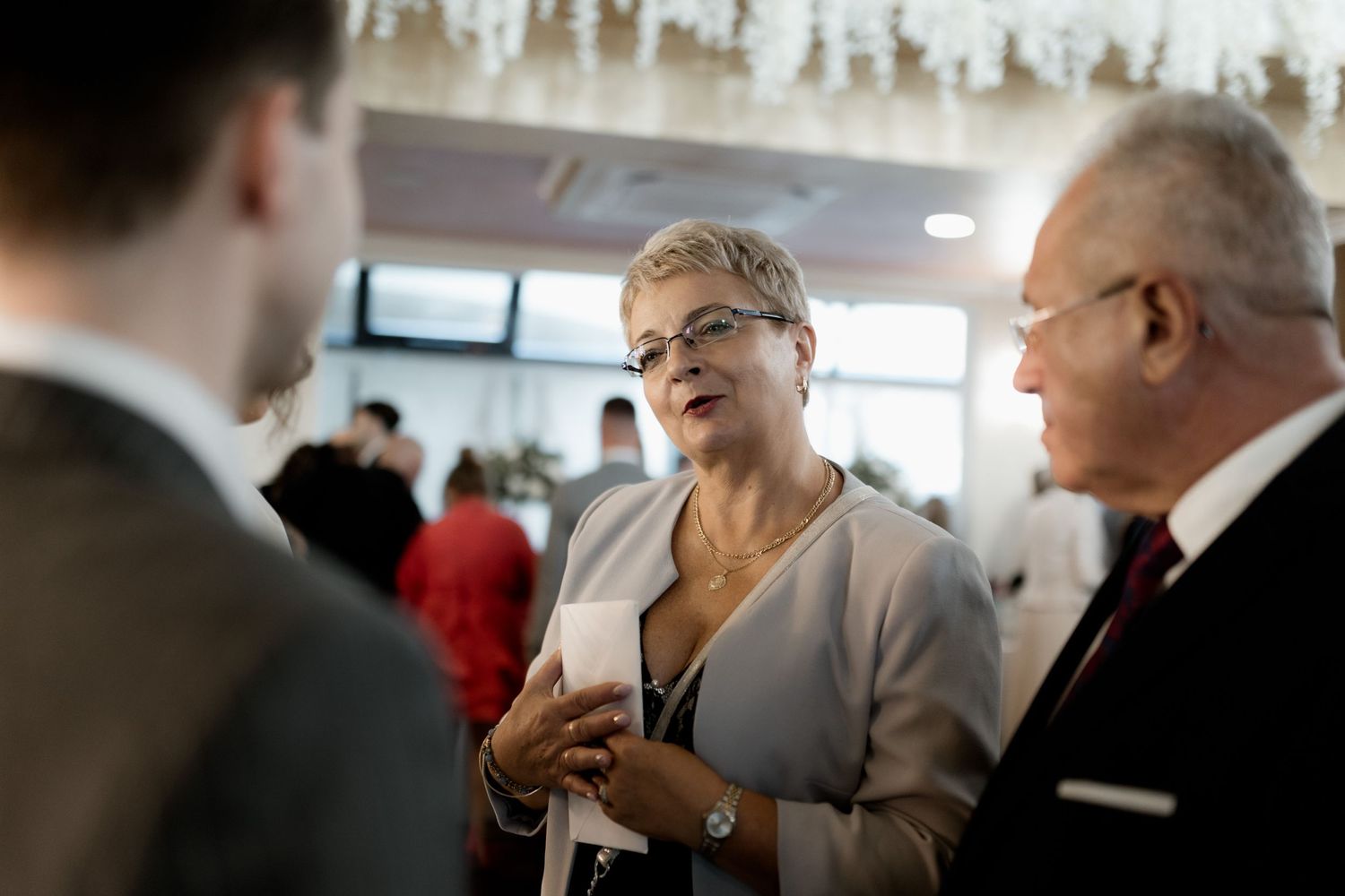 A woman in a beige blazer smiles during a conversation with two people at a professional gathering.
