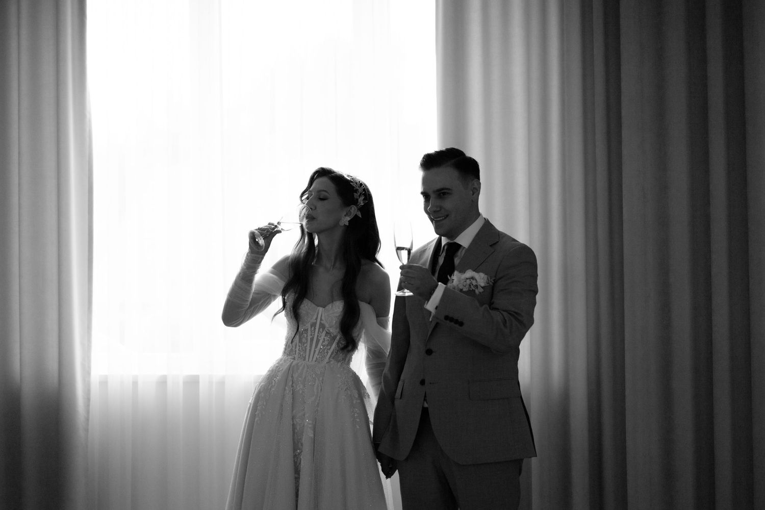 Black and white photo of a couple drinking from champagne glasses in a bright room with curtain backdrop.