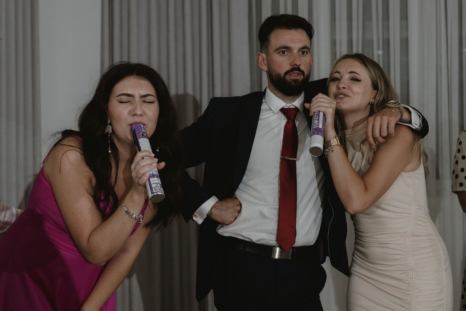 Three people at a party pose together while drinking beverages in front of a curtain backdrop.