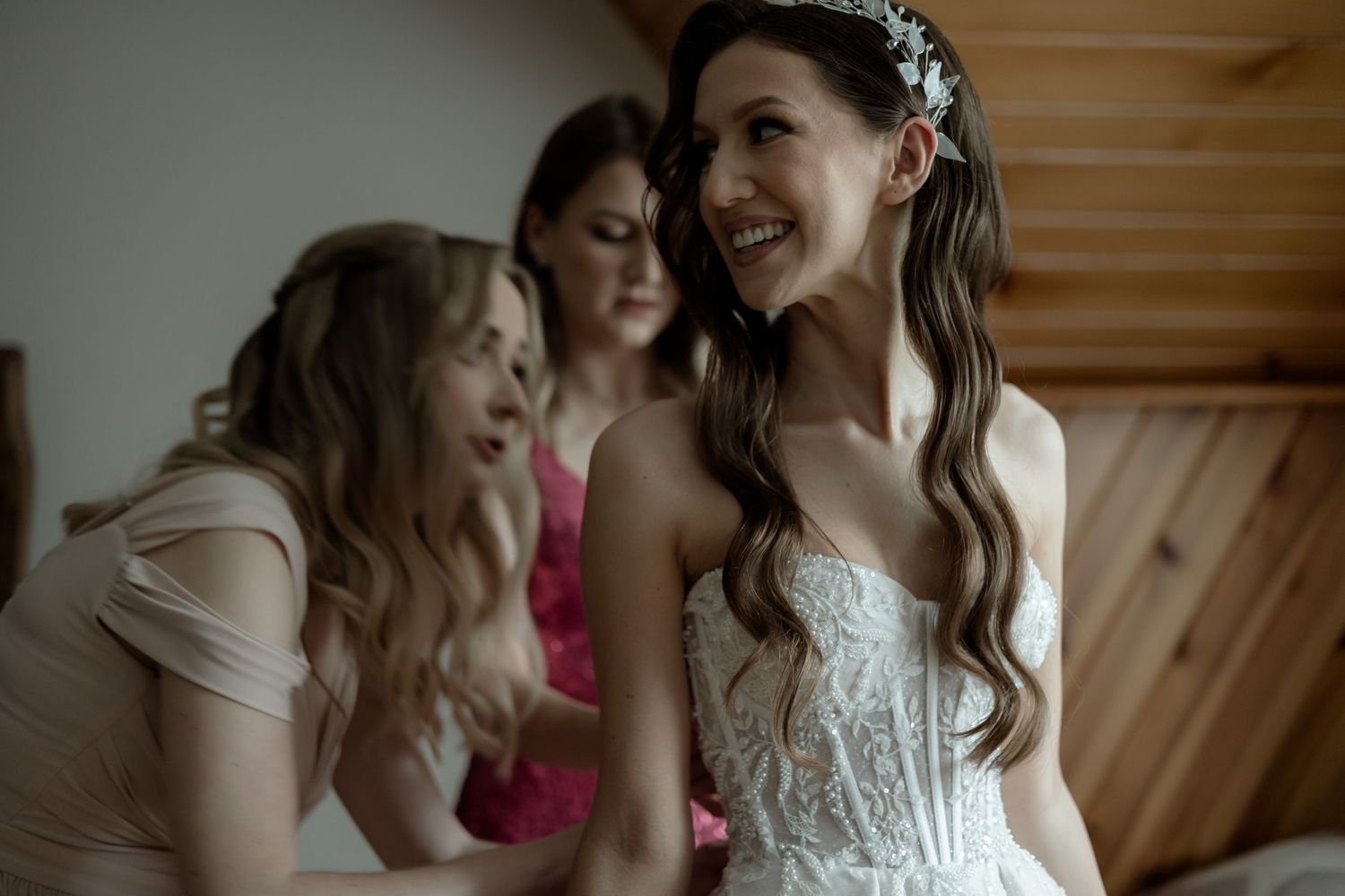 Bride in white lace corset dress shares joyful moment with bridesmaids in blush gowns at rustic venue.