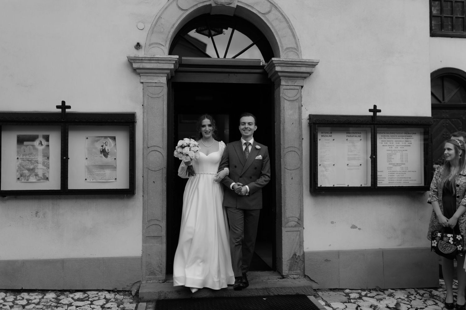 A bride and groom standing in an ornate stone doorway entrance with vintage movie posters on the wall outside.