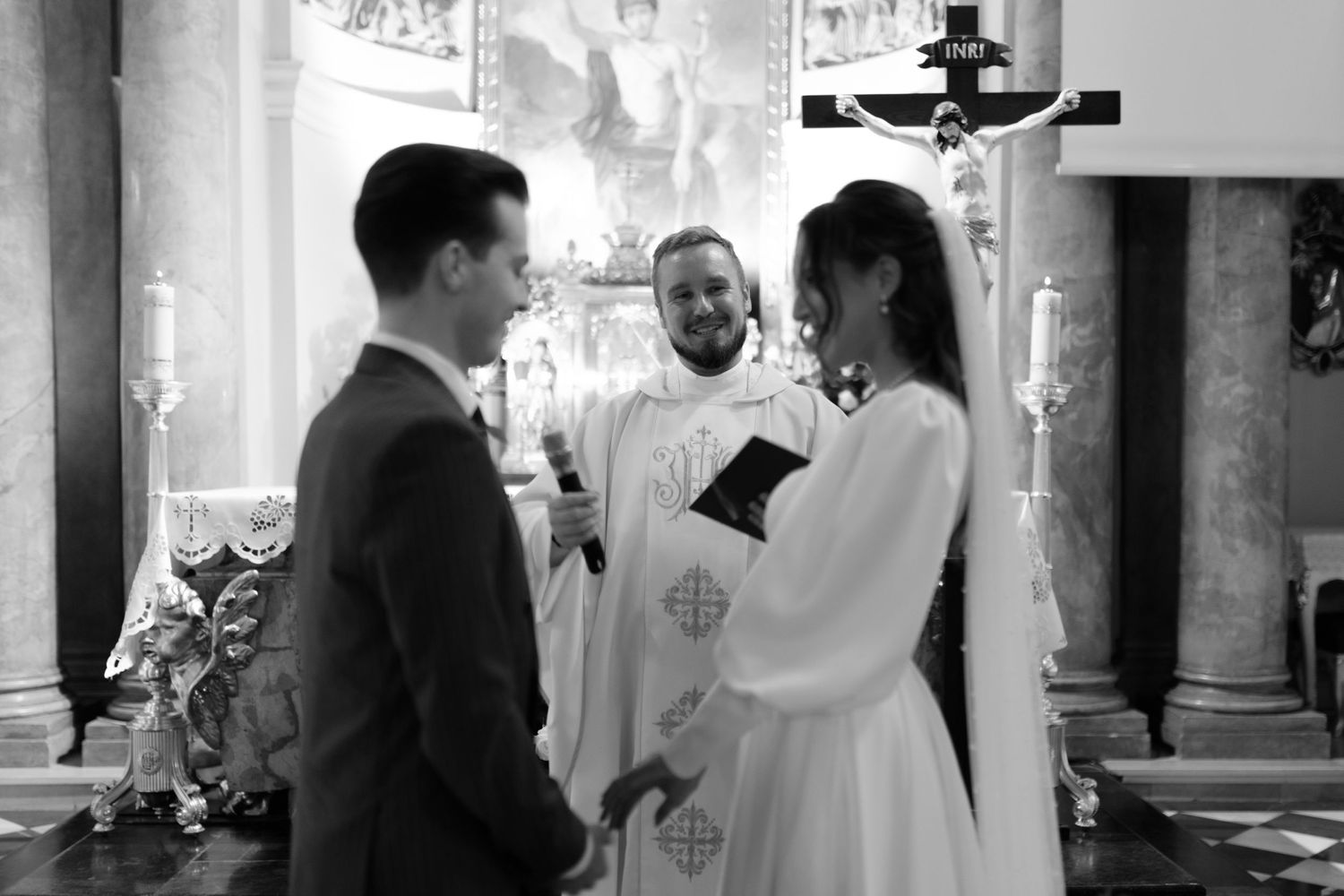 A black and white photograph of a couple during their wedding ceremony with a priest conducting the service.