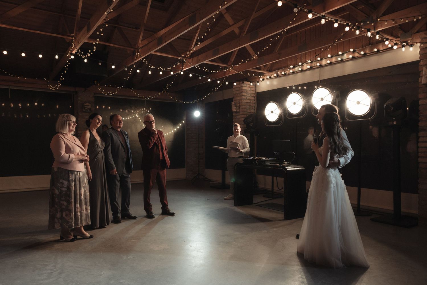 A couple shares their first dance at a rustic indoor venue with string lights and round paper lanterns overhead.
