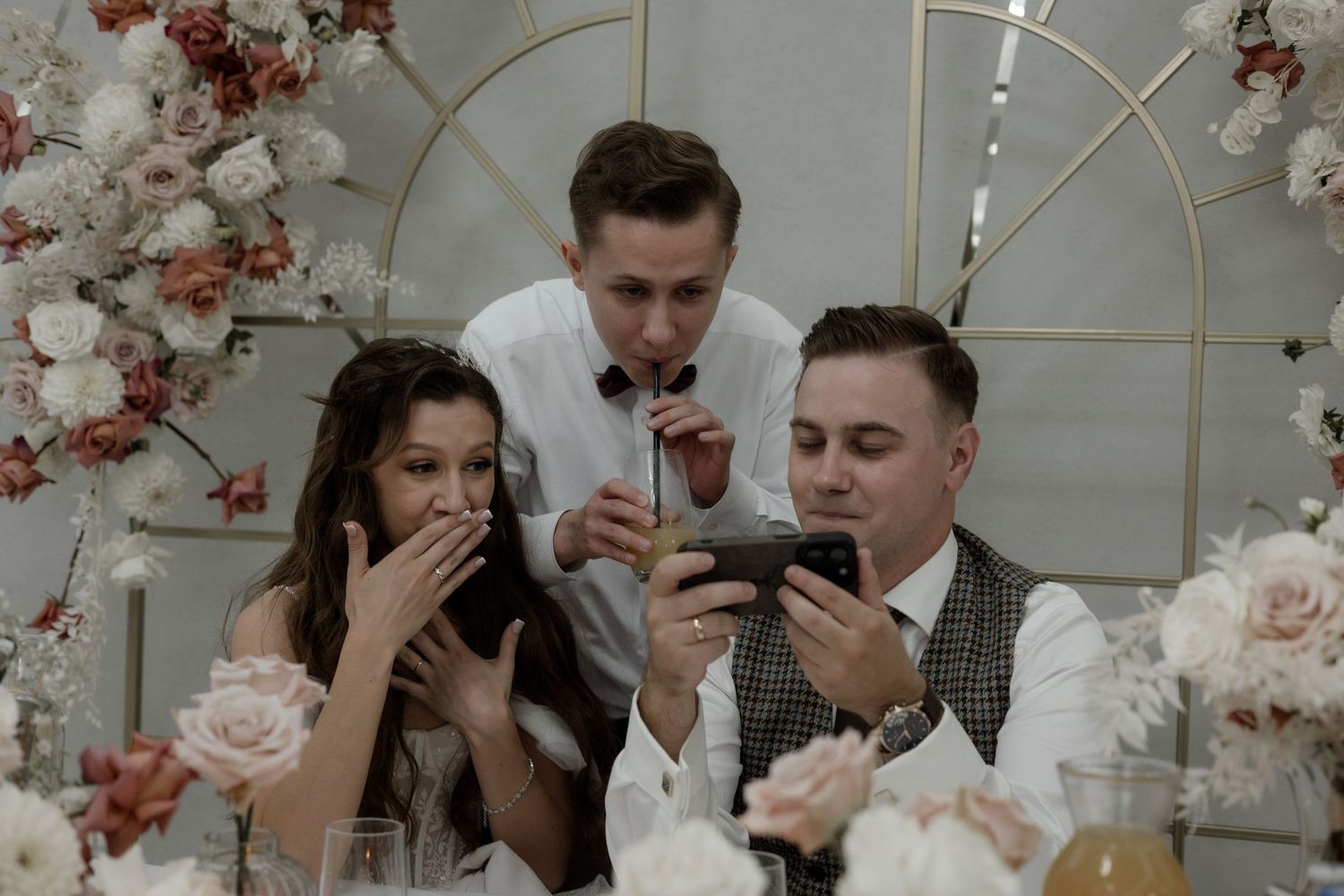Three wedding guests look at their phones with surprised expressions in a floral-decorated venue setting.