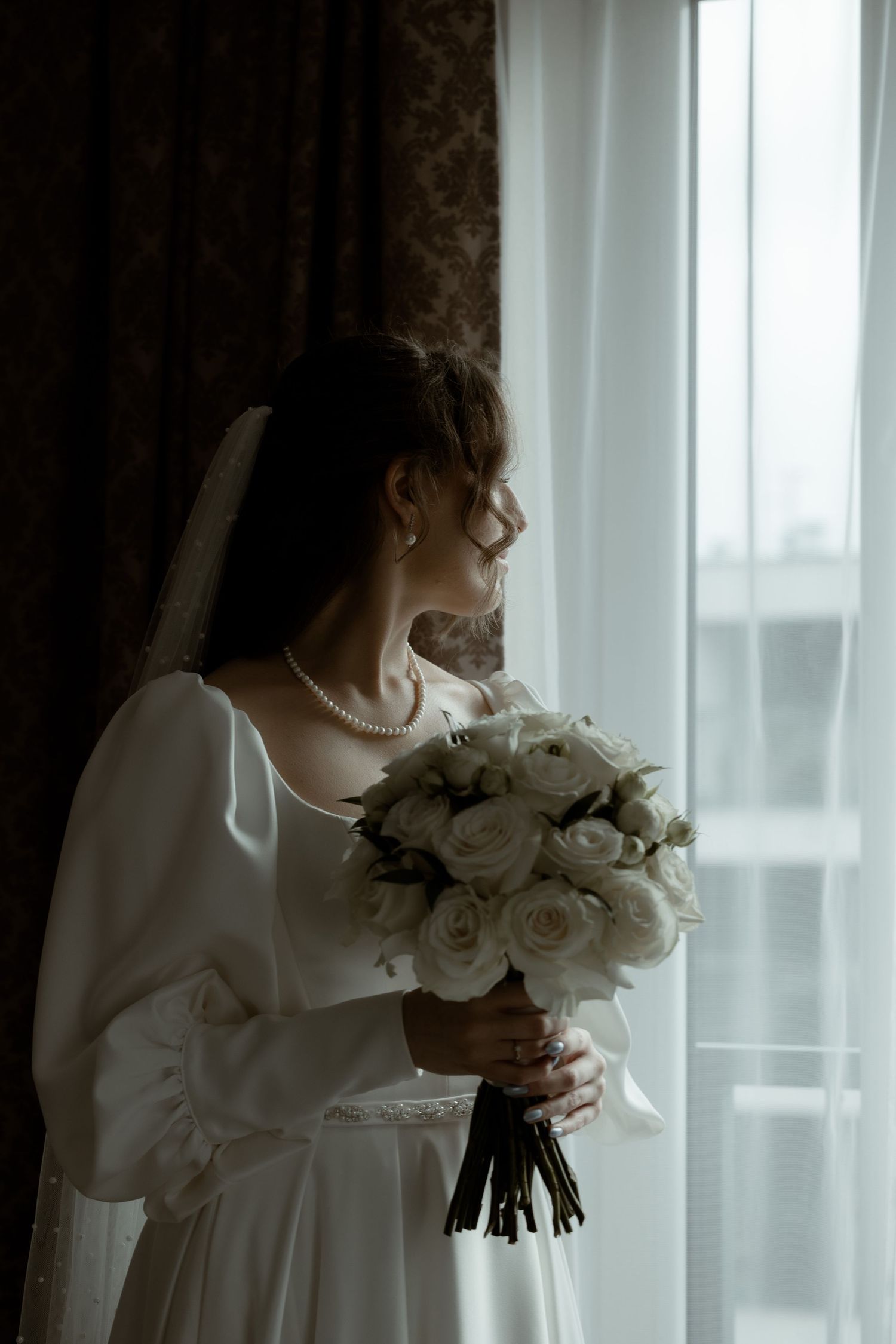 Side profile of bride in elegant white dress holding beautiful white rose bouquet by window in moody lighting.