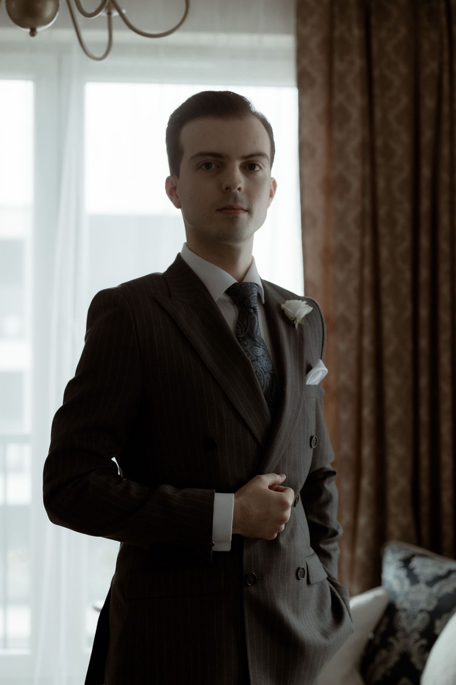 A person wearing a stylish dark suit and tie stands in a room with brown curtains and natural window lighting.