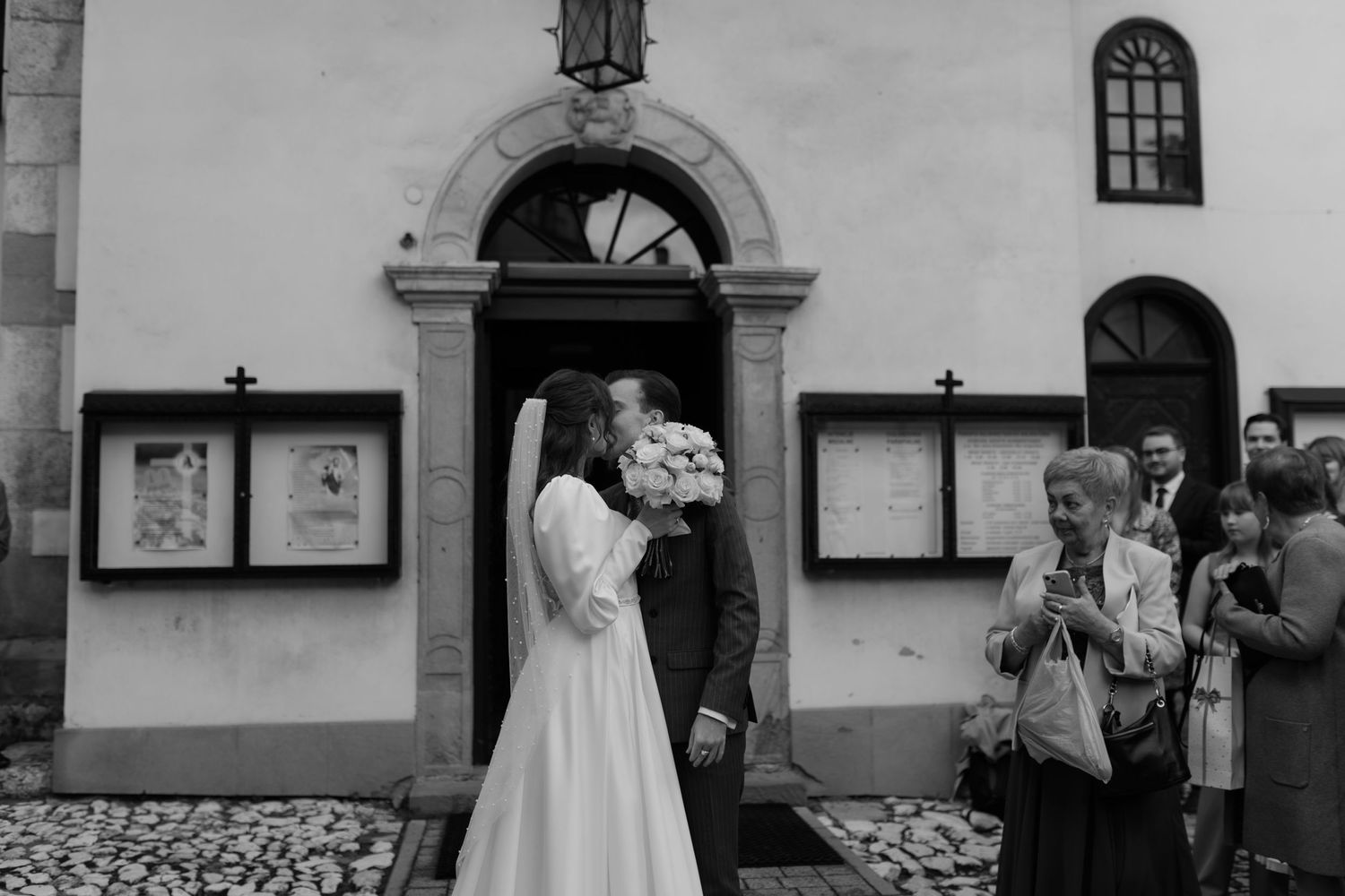 Black and white photograph of a wedding moment outside a historic church with arched doorway and display cases on the wall.
