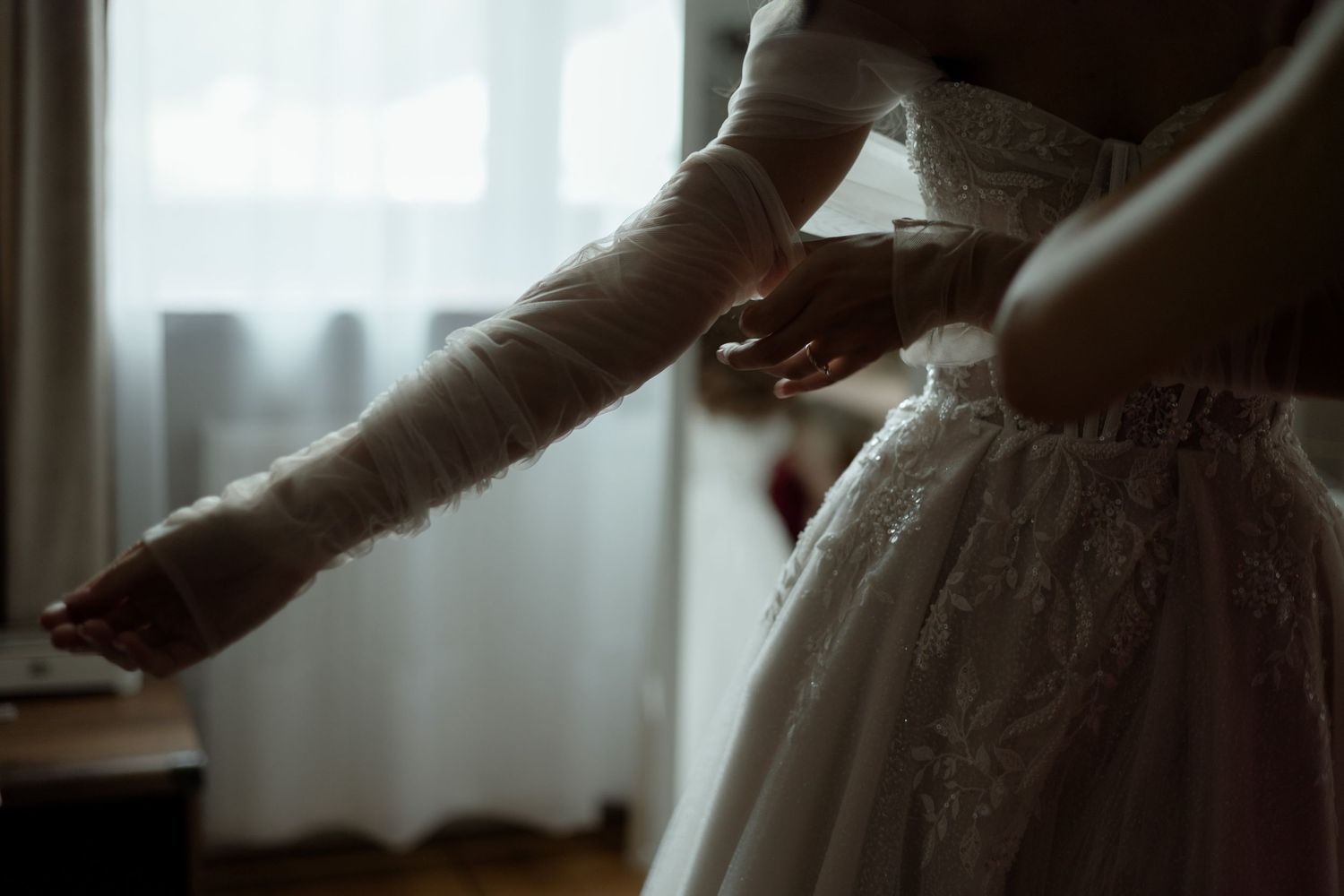 An elegant white wedding dress with delicate lace details being adjusted in soft natural window light.