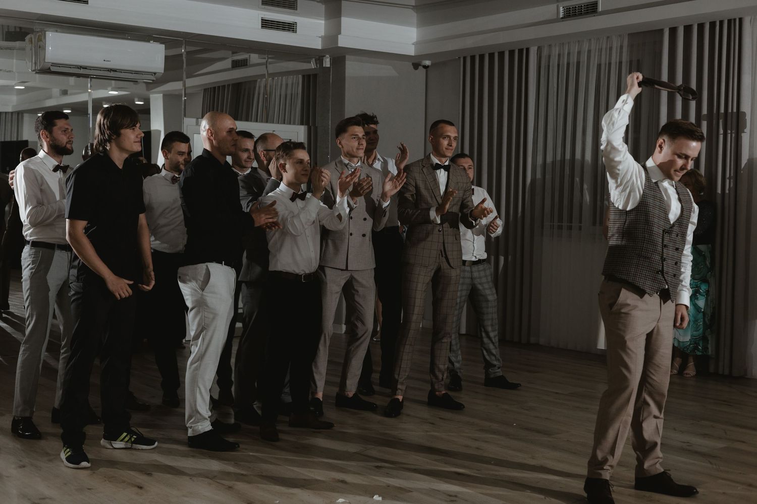 A group of wedding guests and groomsmen celebrate together on a dance floor during a reception.