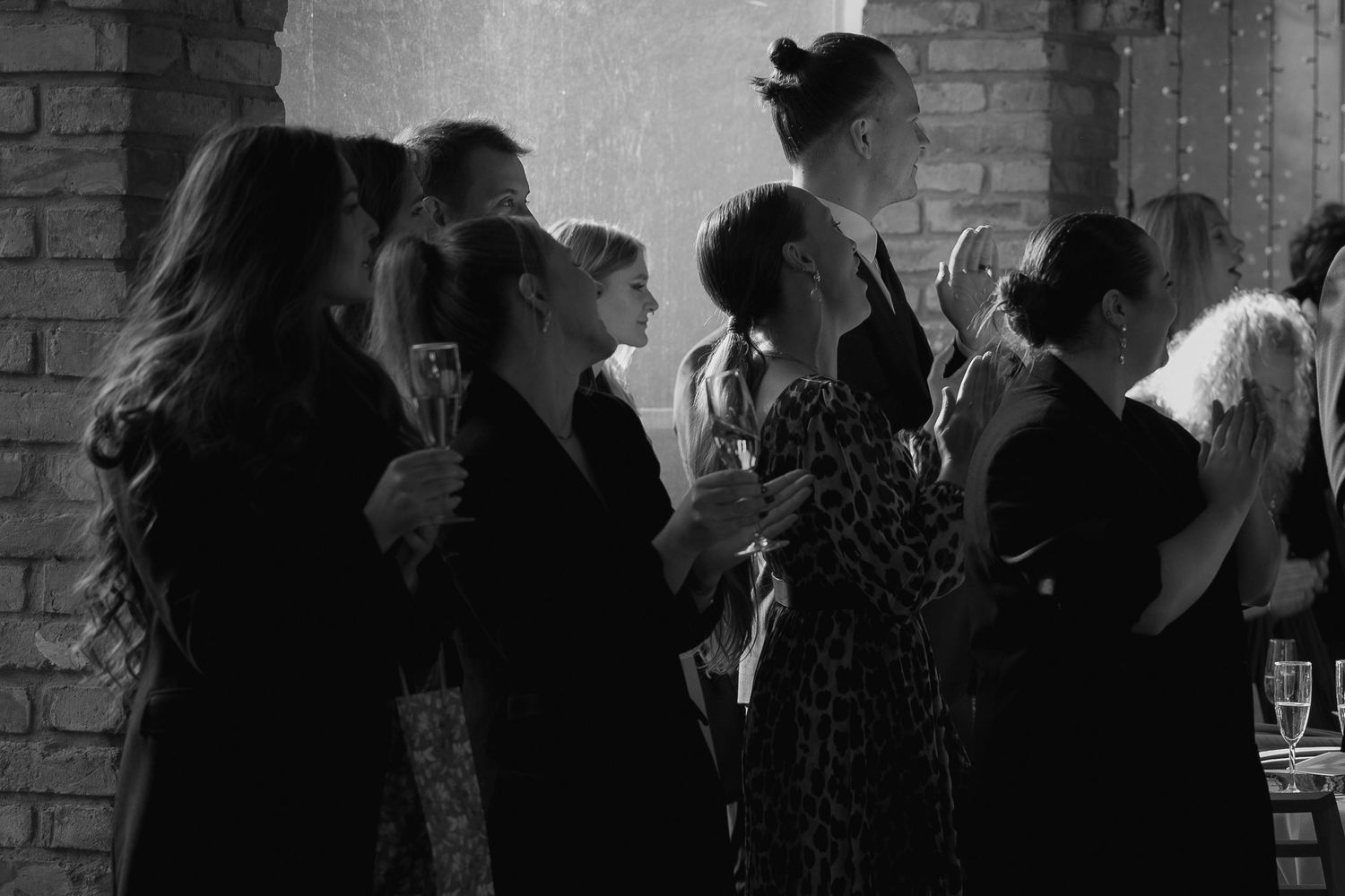 A black and white photograph shows a group of people standing together in silhouette against a brick wall at a social gathering.