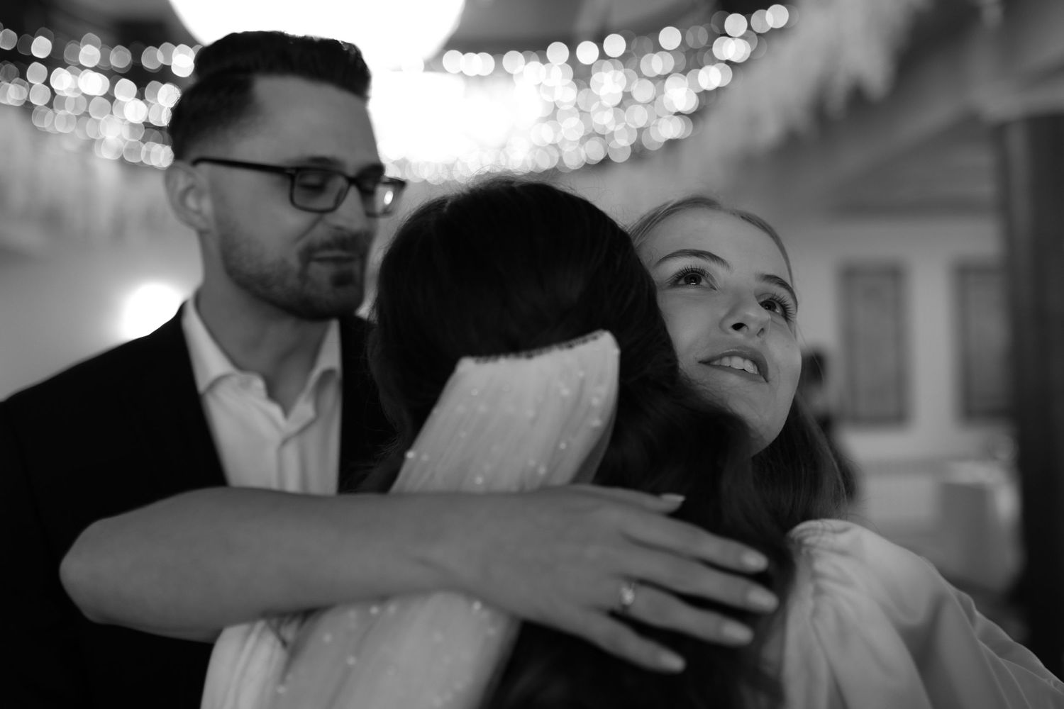 Black and white photo of an embracing group at an indoor event with bokeh lighting in the background.