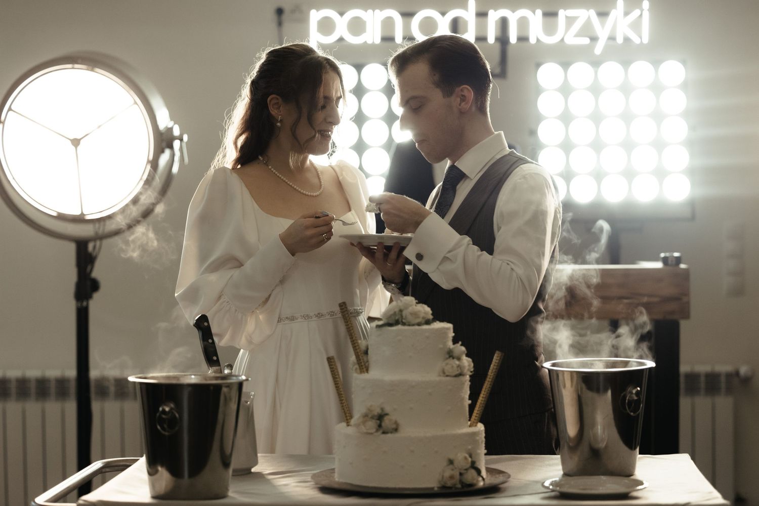 Elegant couple in formal attire sharing a romantic moment while cutting cake in front of bright studio lights.