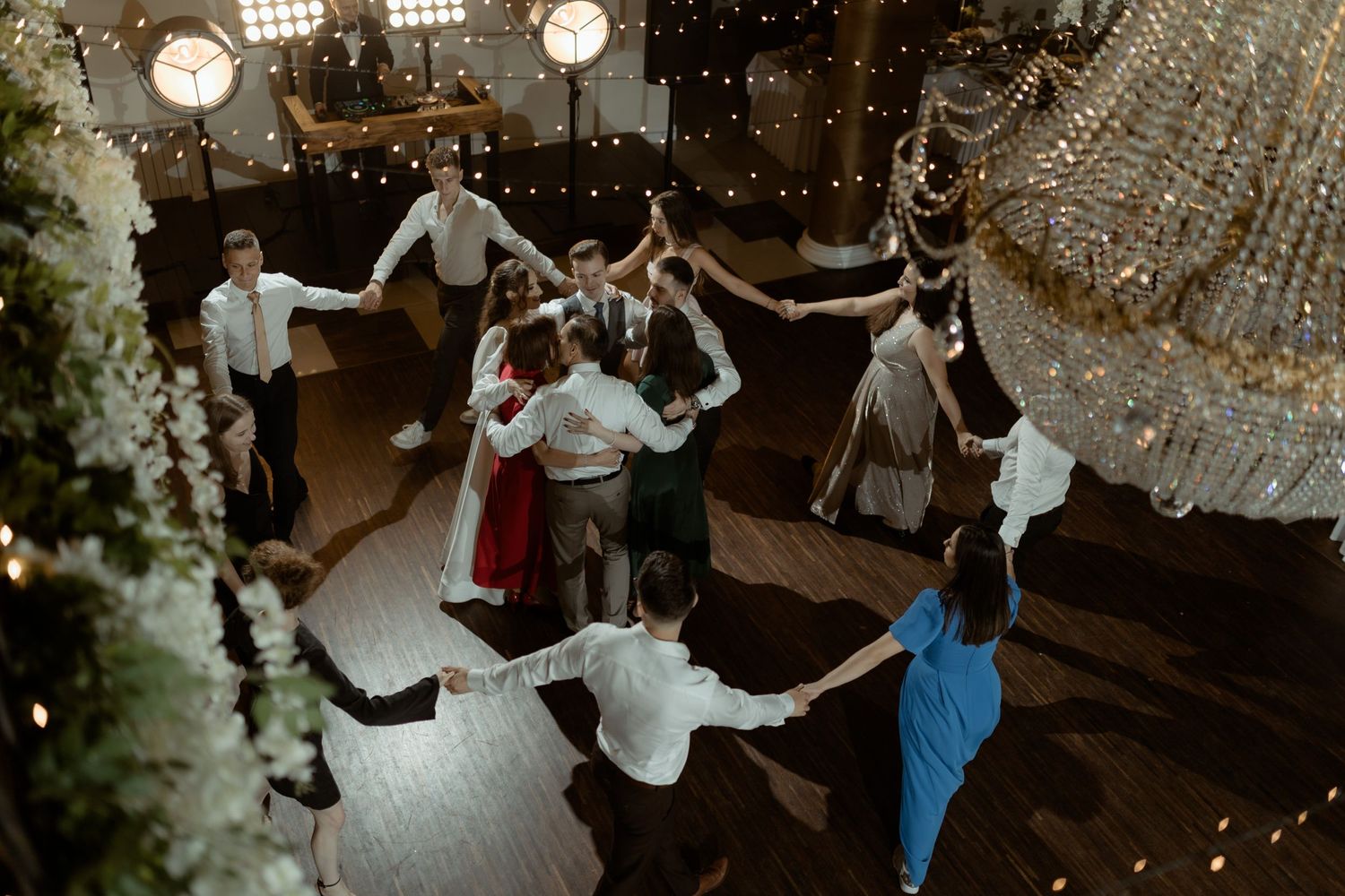 Wedding guests dance in a circle under twinkling lights and chandeliers during a festive reception celebration.
