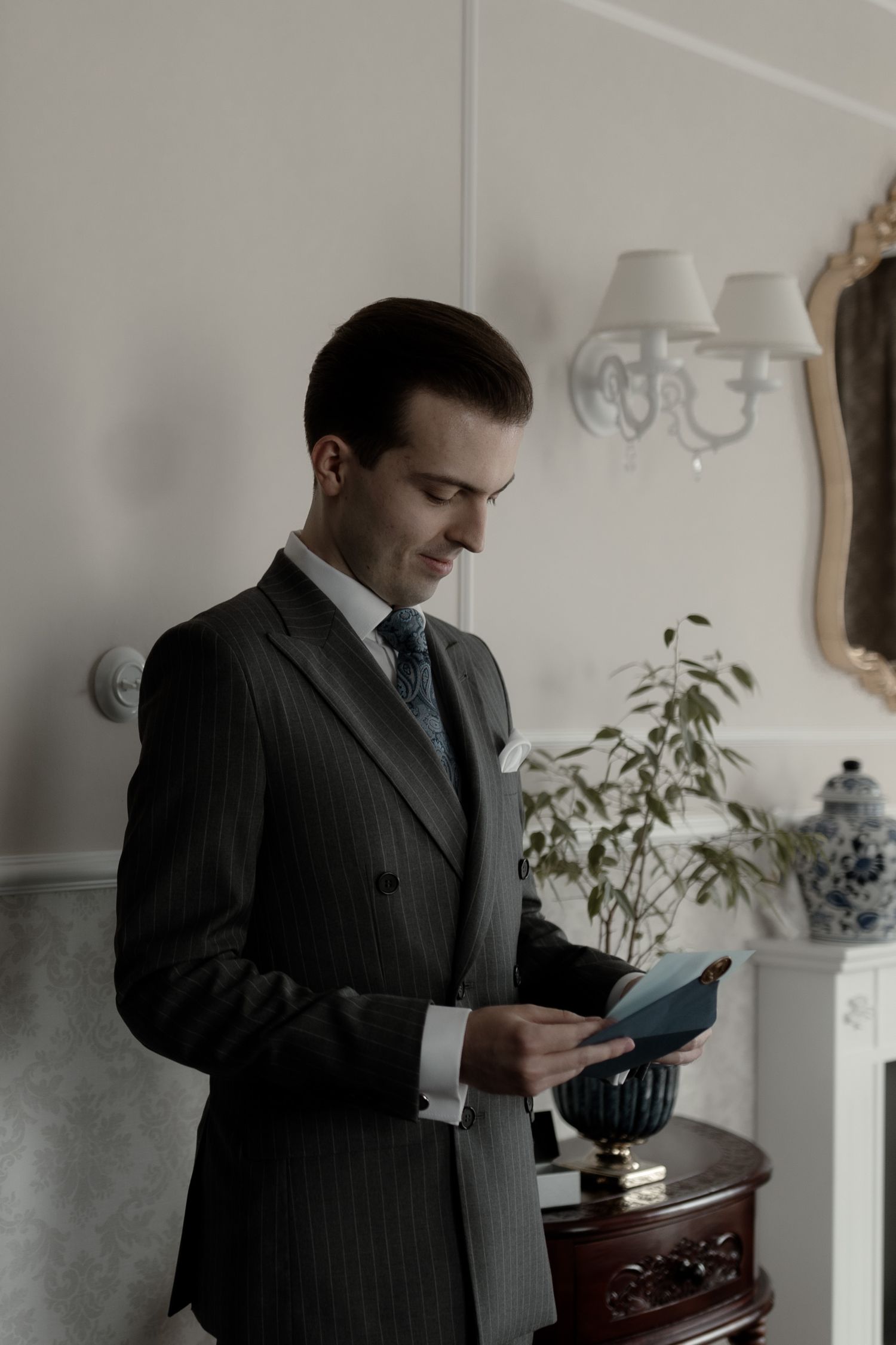 A person in a pinstripe suit looks down at a mobile device while standing in an elegant room with wall sconces.