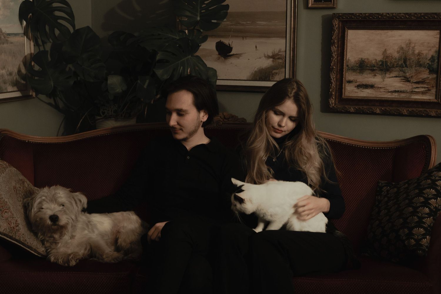 A couple sits on a vintage couch with two pets - a white cat and a small dog in a dimly lit room.