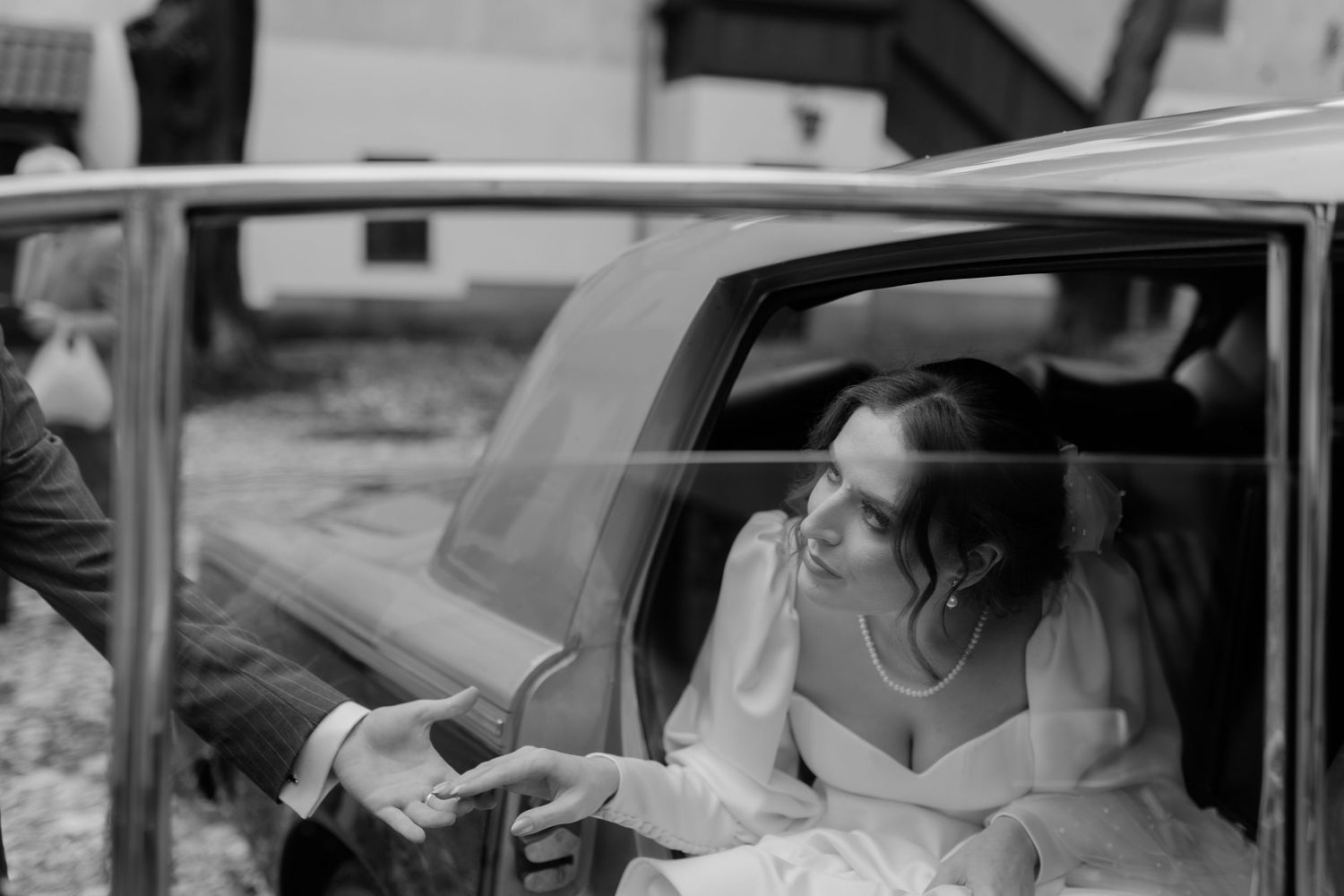 A bride in a wedding dress reaches out while seated in the backseat of a vintage car during a black and white moment.