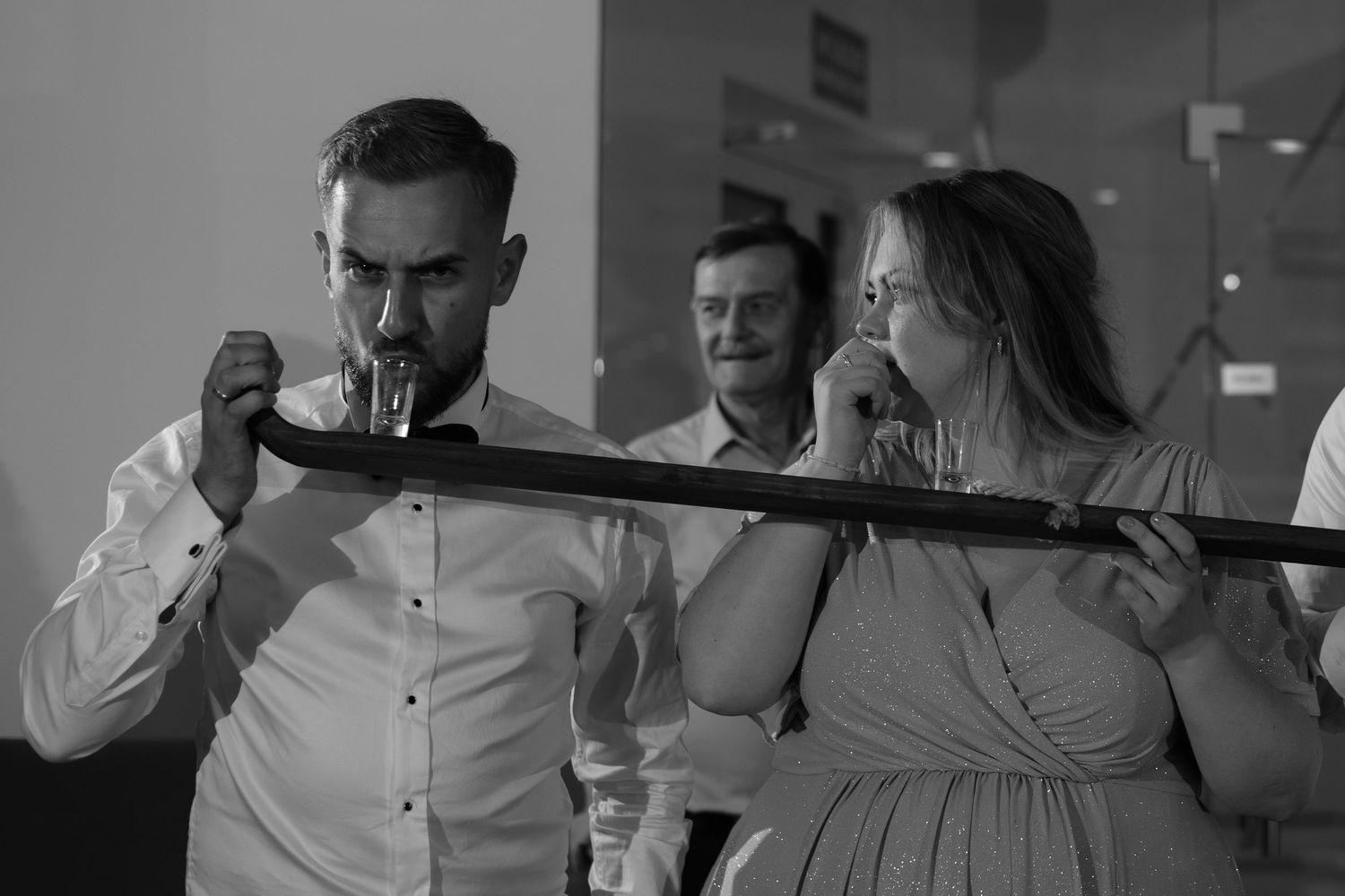 A couple sharing a drink from a sword at what appears to be a wedding reception or party, photographed in black and white.