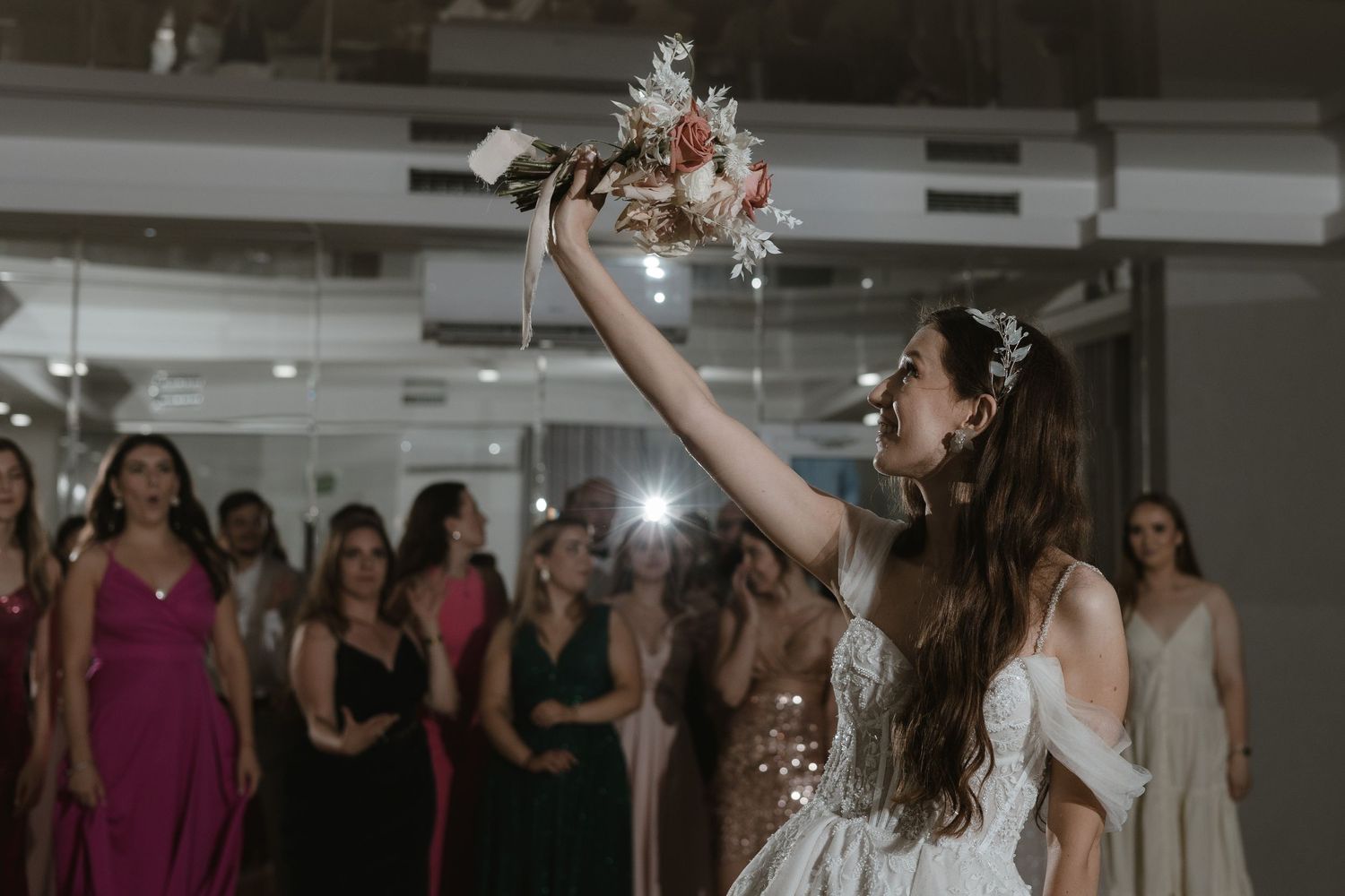 A bride in a white dress raises her bouquet during a wedding reception as guests in formal attire watch from behind.