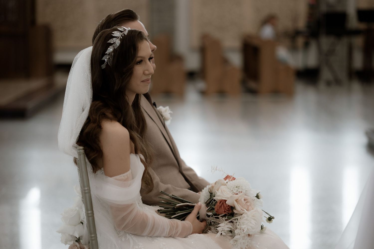 A bride in a white wedding dress sits holding a pink and white bouquet in a soft-lit setting.