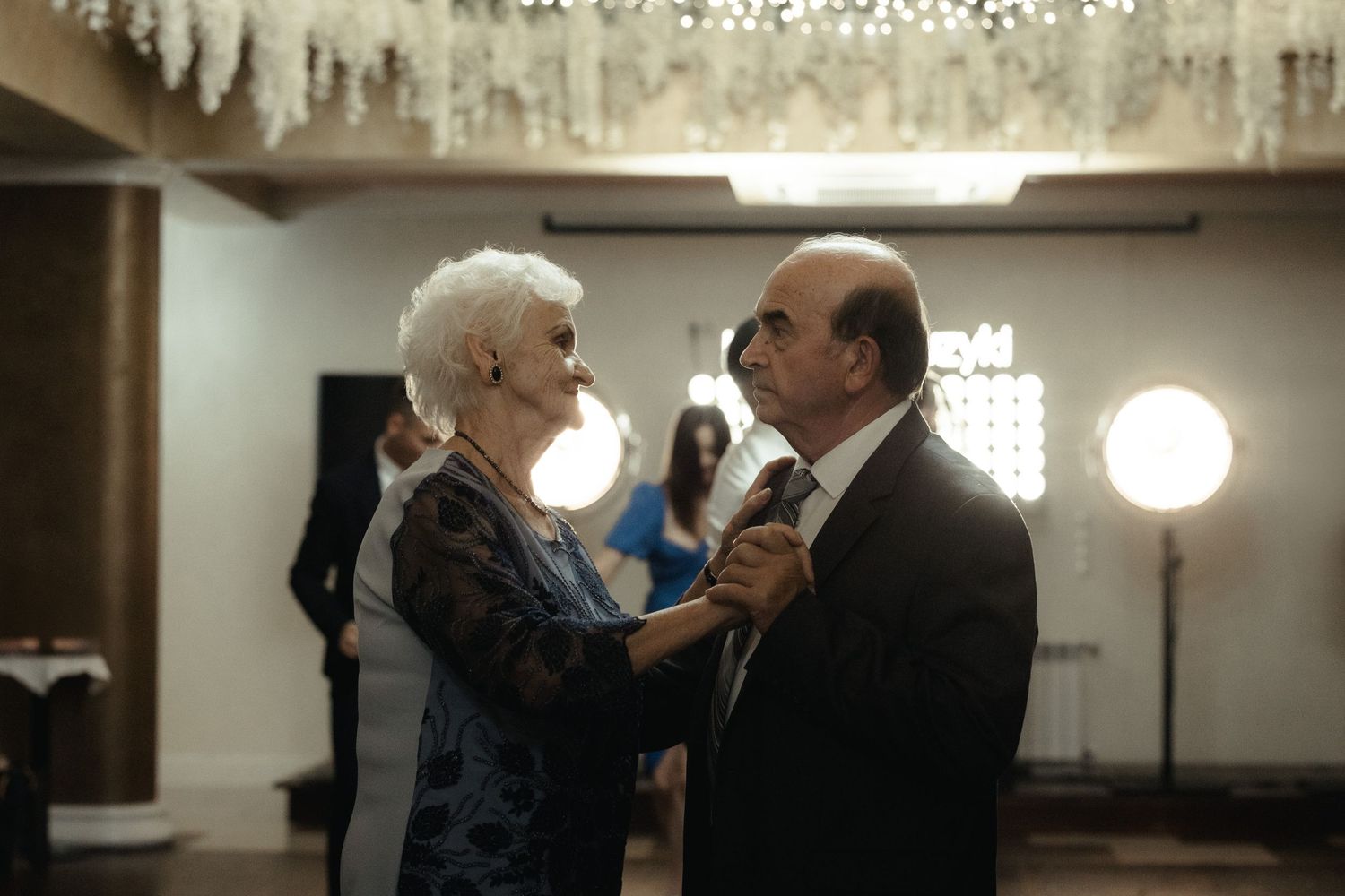 An elderly couple in formal attire sharing a romantic dance moment together in a dimly lit ballroom.