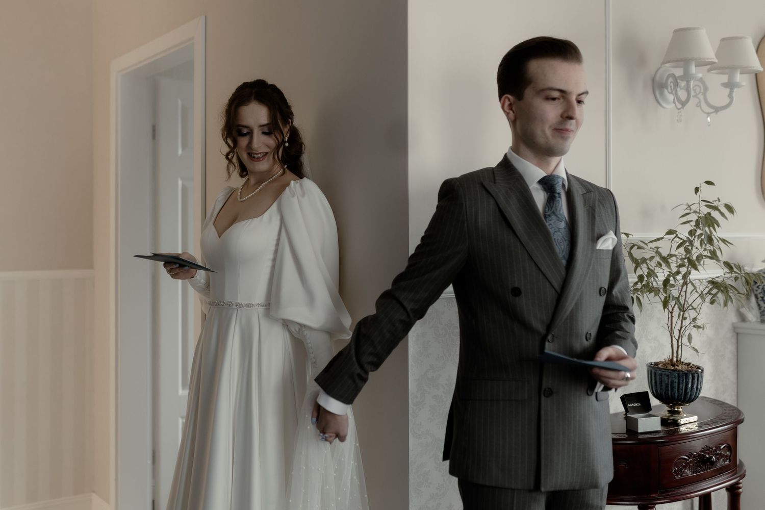 A wedding couple holds hands around a corner without seeing each other in an elegant indoor setting.