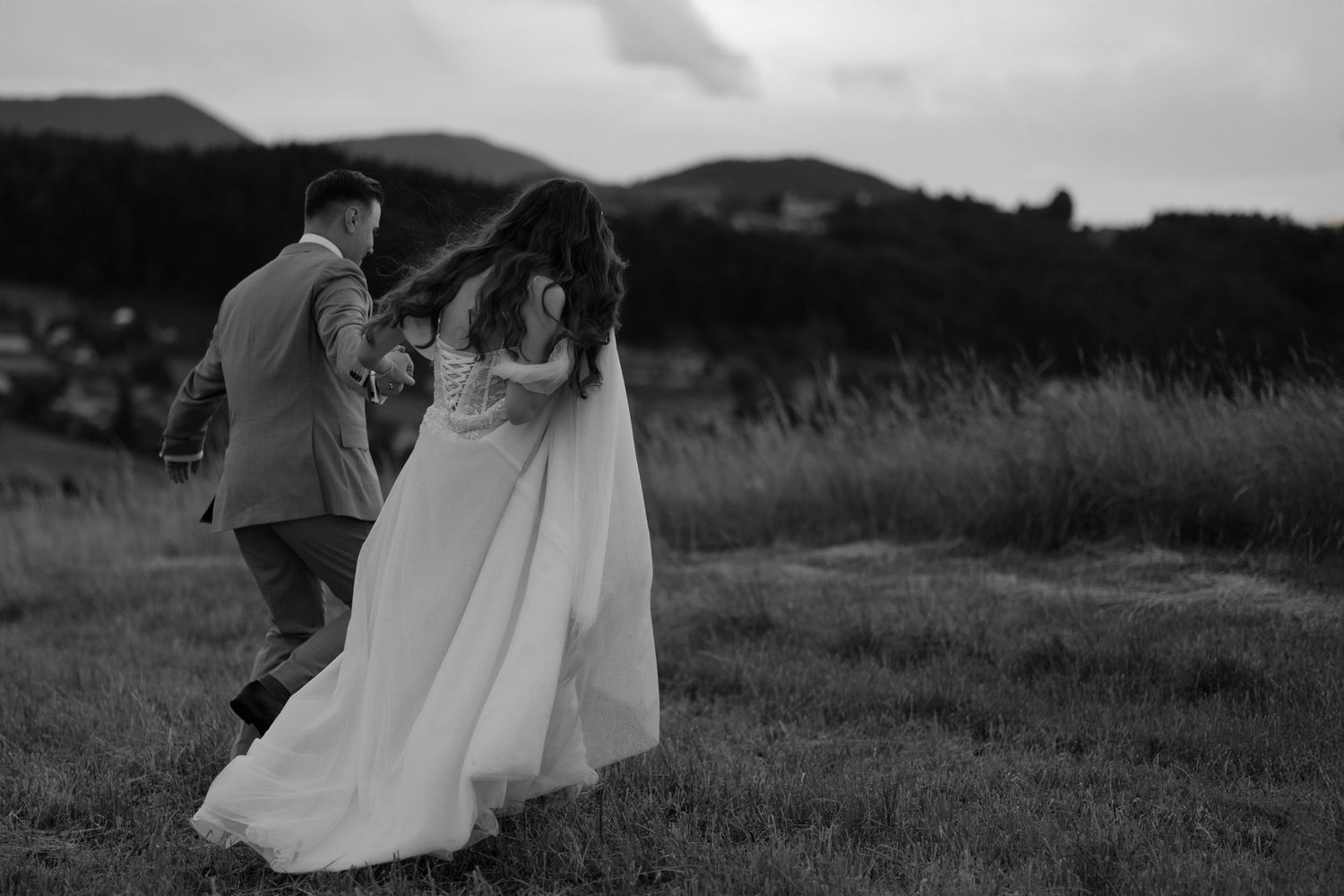 Black and white wedding photo of couple dancing together in a field with mountains in the background at dusk.