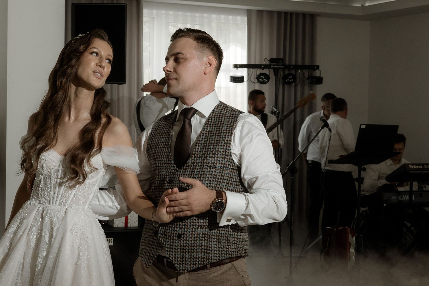 A couple in wedding attire stands together at a reception venue while guests mingle in the background.