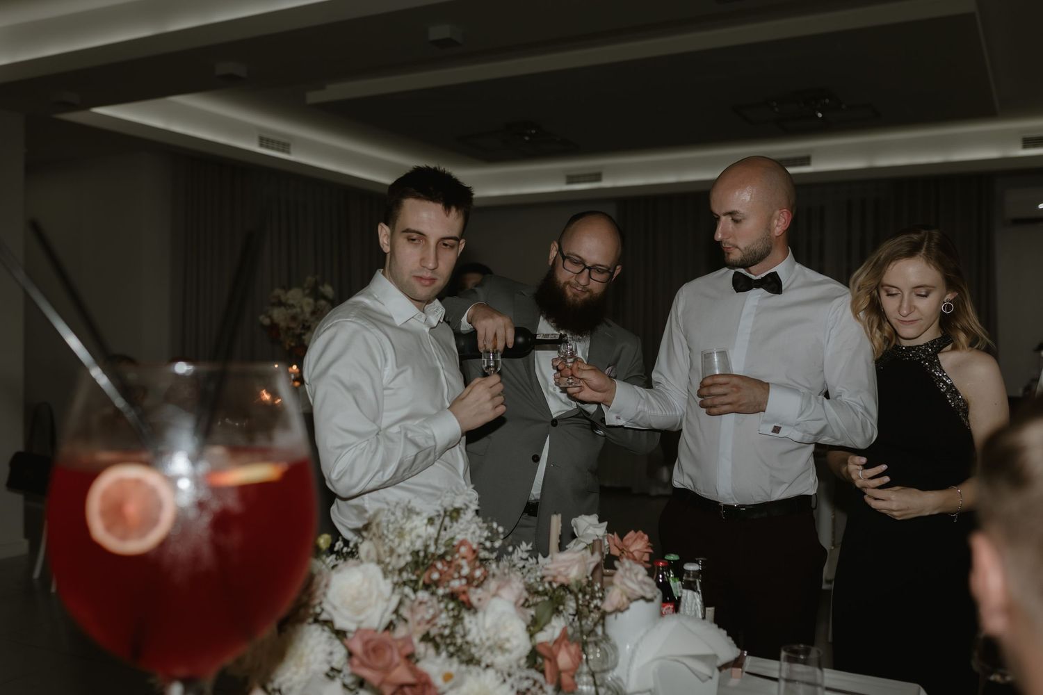 Wedding guests in formal attire gather around looking at their phones during an evening reception.