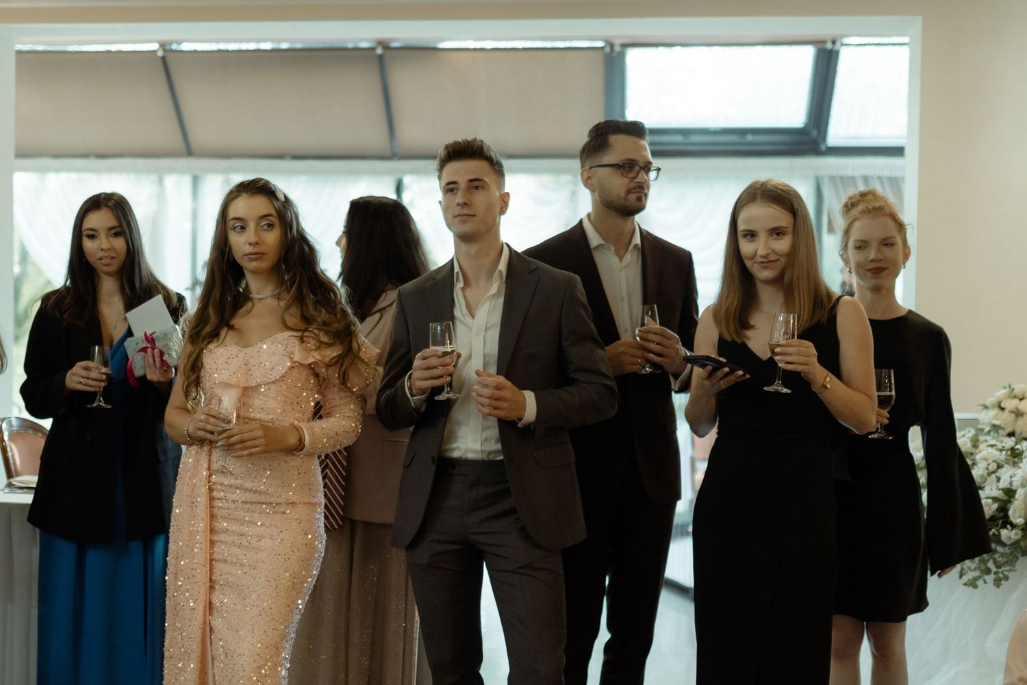 A group of formally dressed guests holding drinks during an upscale social gathering in a bright indoor space.