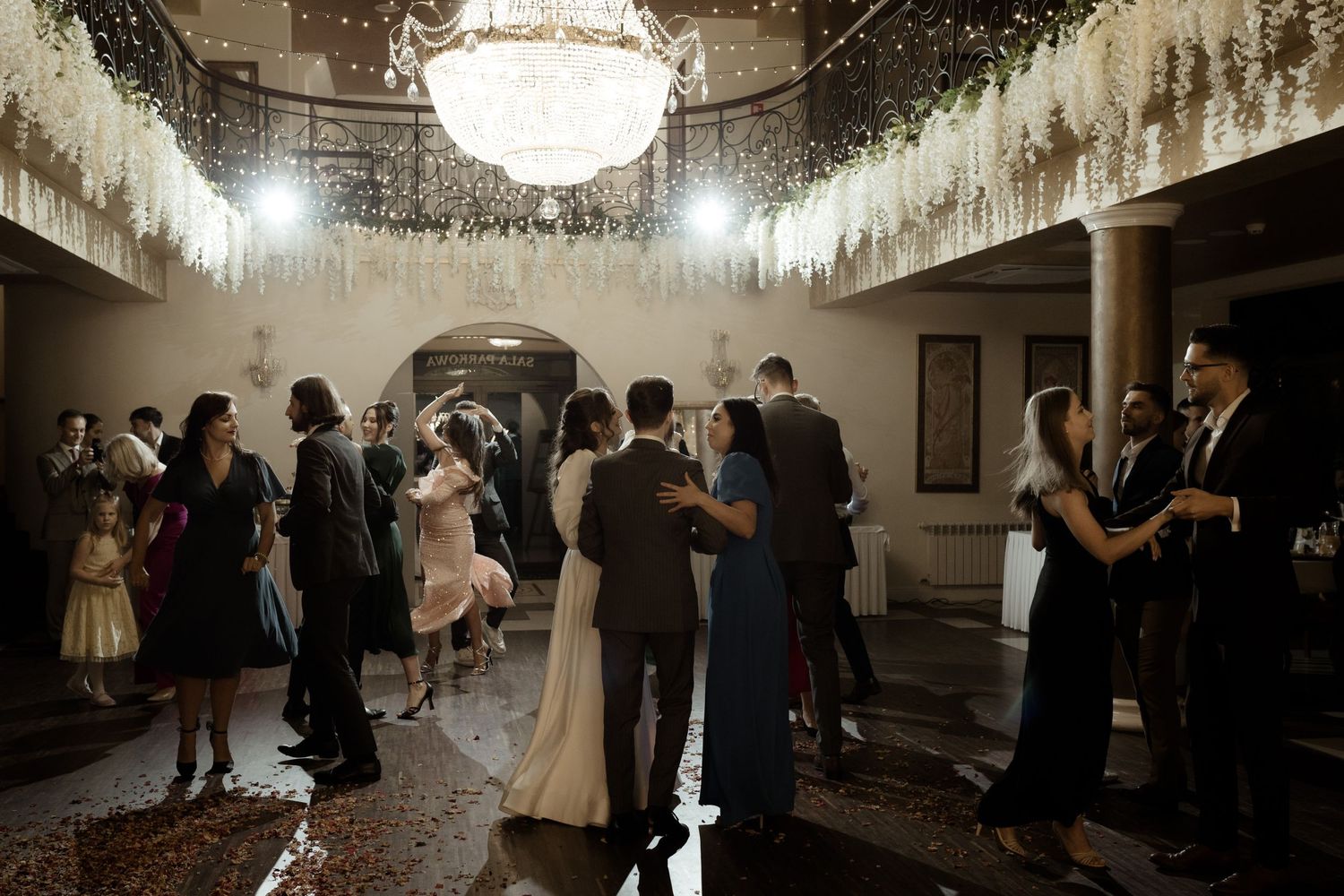 Elegantly dressed guests dancing at a formal reception under a grand chandelier in a dimly lit ballroom.