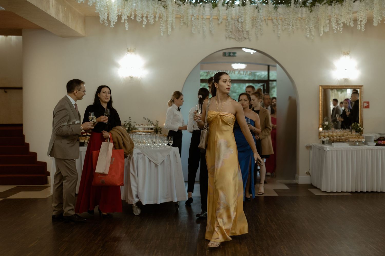 Guests in formal attire gather at an indoor wedding reception venue with hanging white decorations and buffet tables.