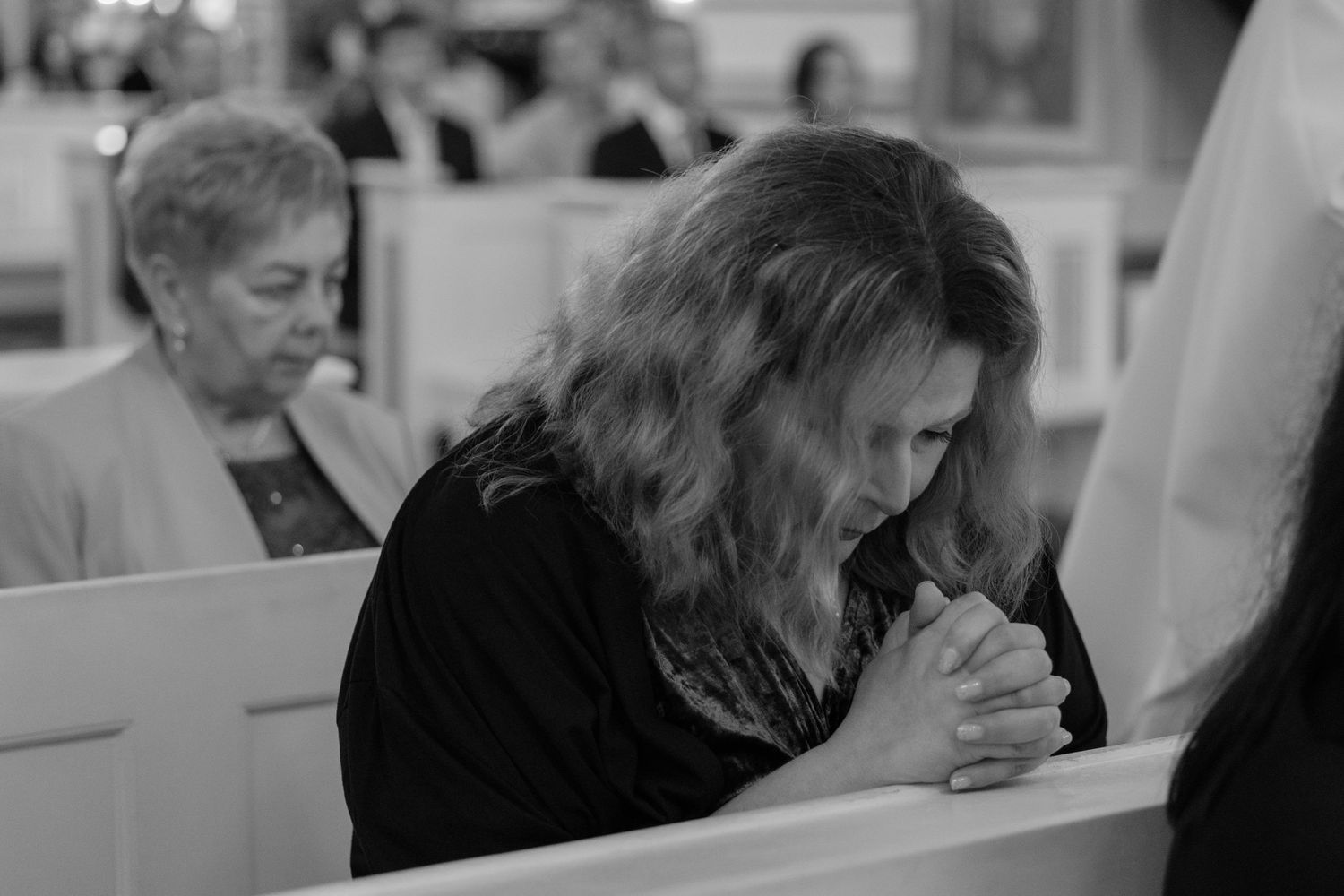 A person in a black sweater bows their head in prayer while sitting in a church pew.