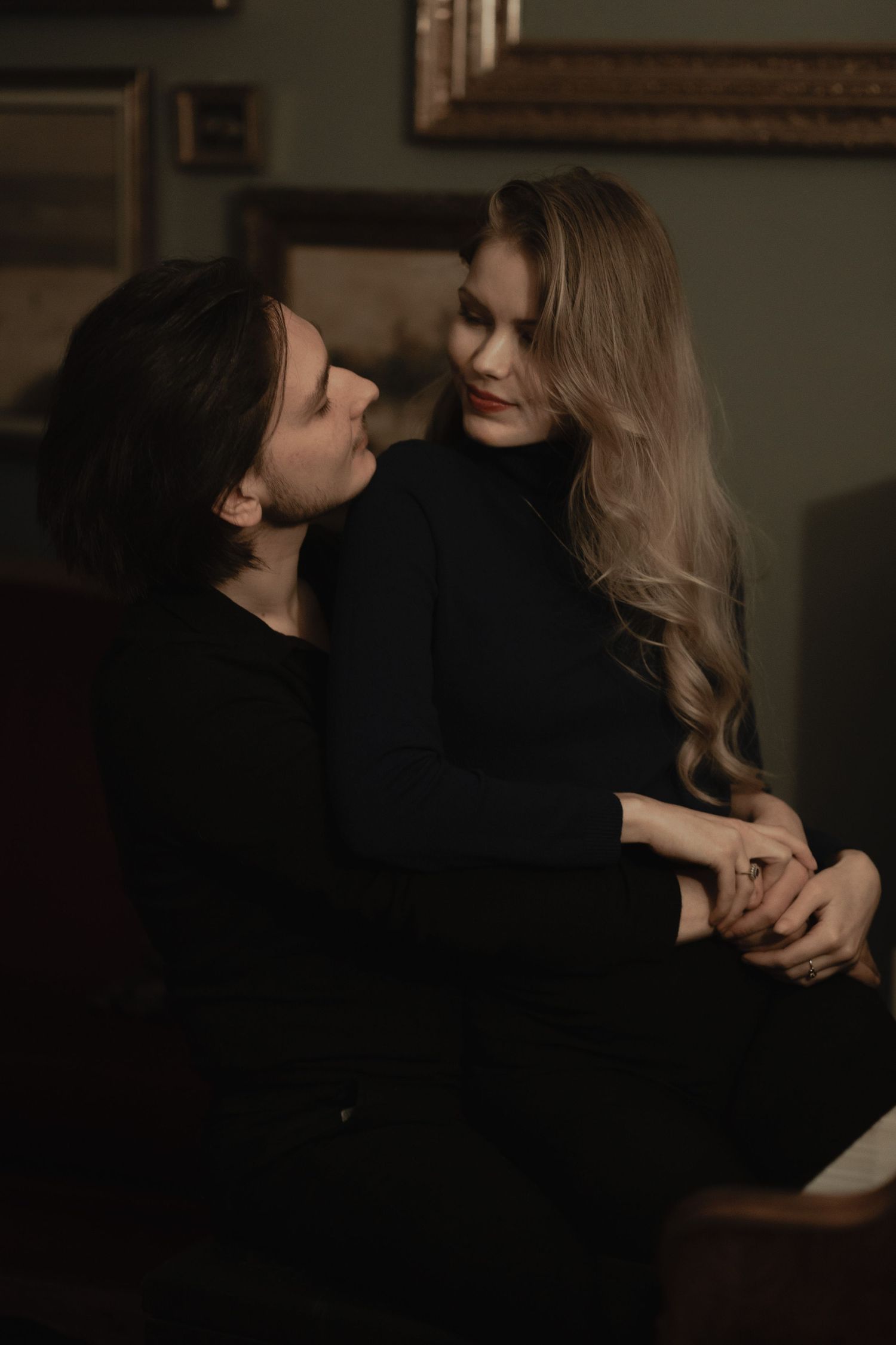 A moody intimate portrait of two people in black clothing sharing a tender moment in dim lighting.