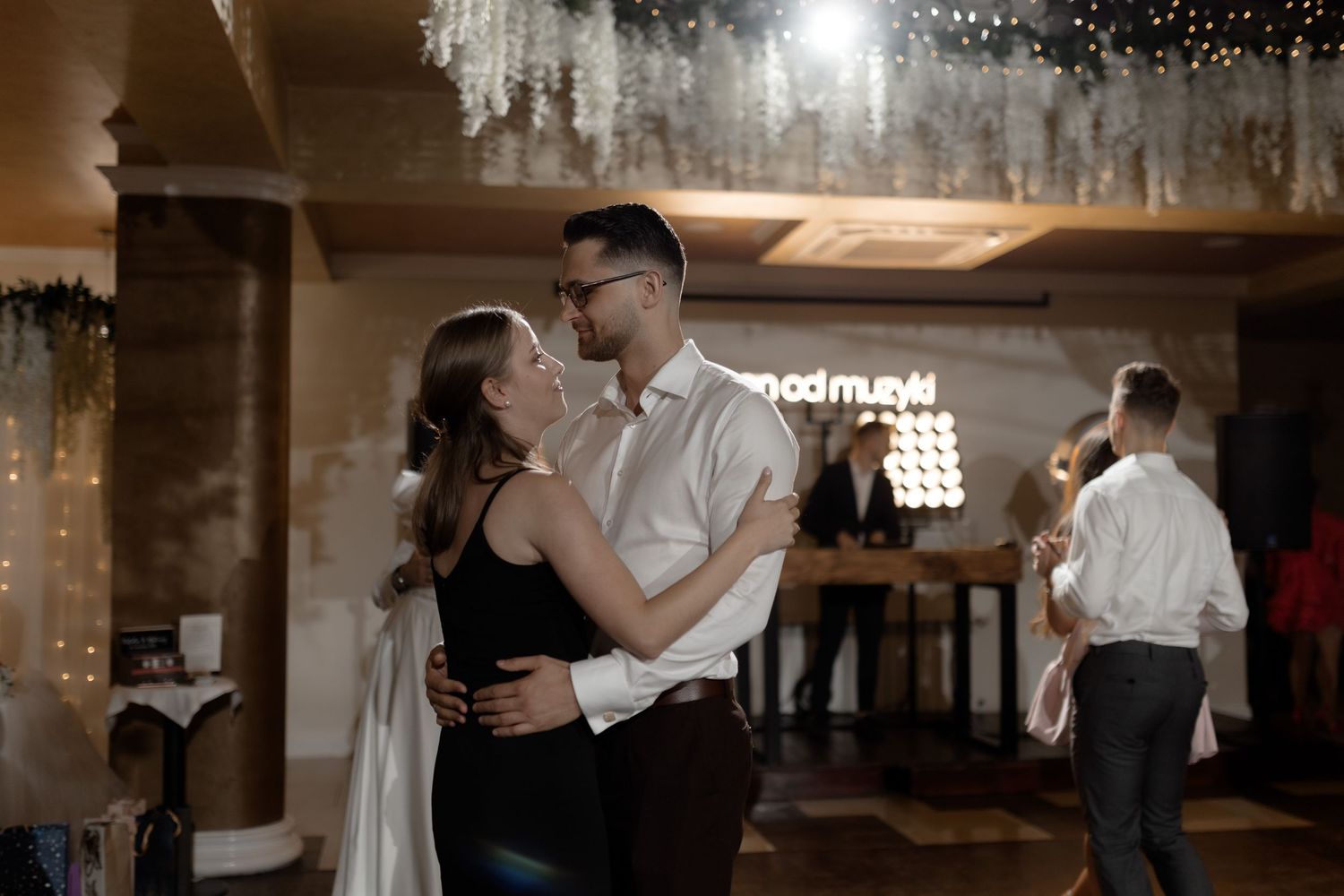 A couple shares an intimate dance moment at an indoor reception venue with twinkling lights in the background.