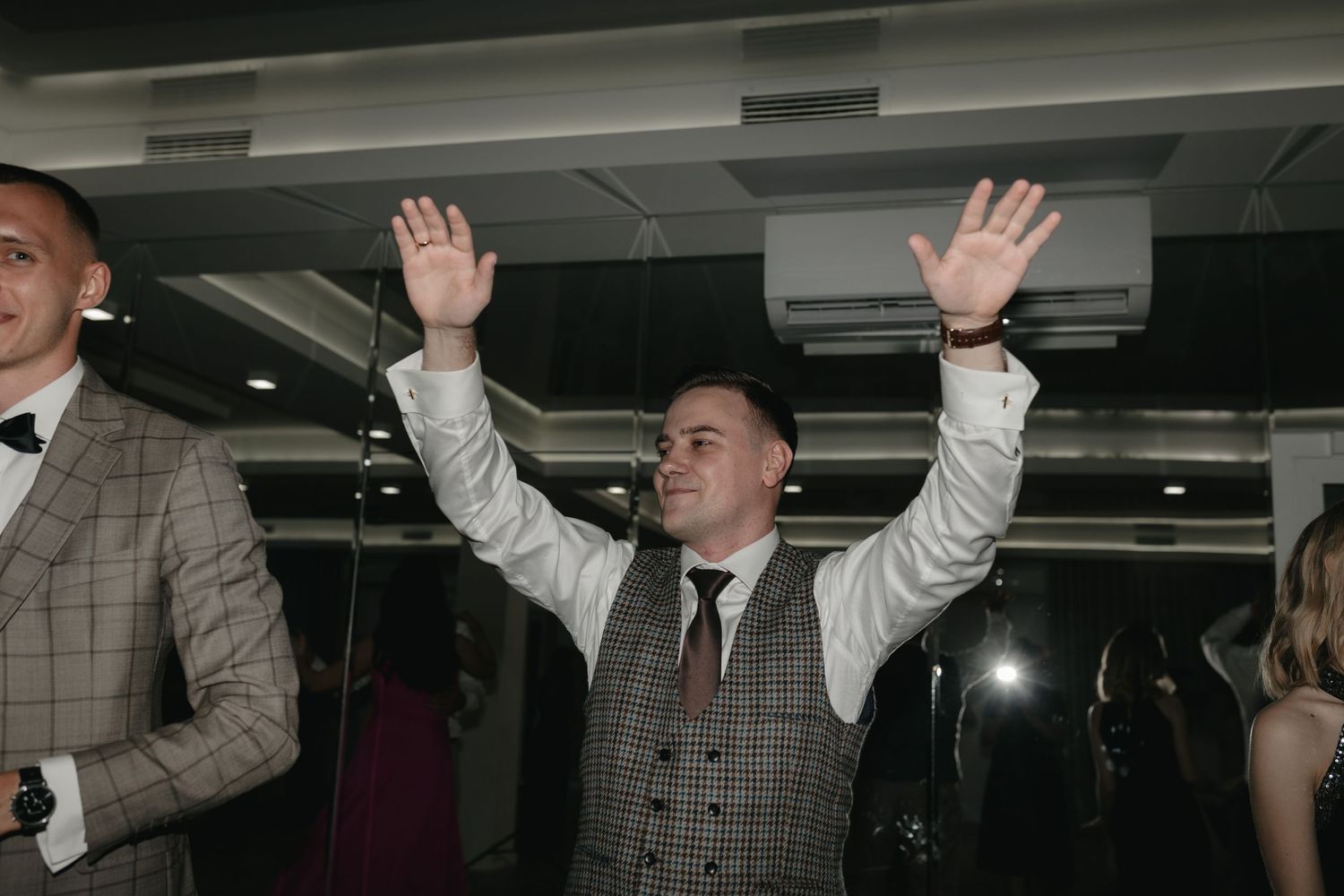 Wedding guest raises arms in celebration while wearing a gray tweed vest and tie during indoor reception celebration.