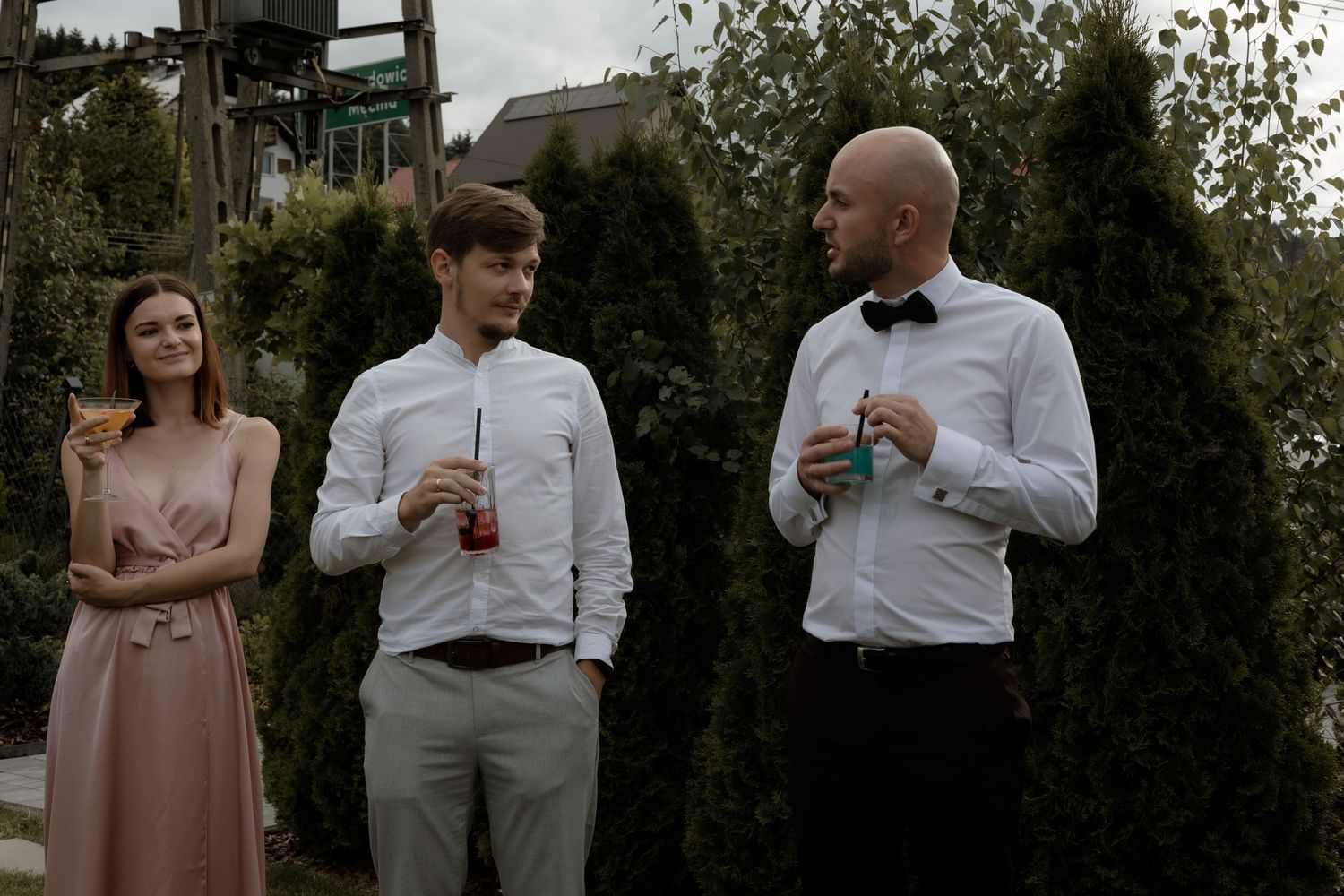 Three wedding guests stand together in formal attire holding drinks during an outdoor garden reception.