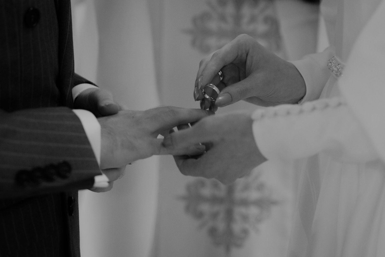A black and white photo of hands exchanging wedding rings during a ceremony in front of a decorative backdrop.
