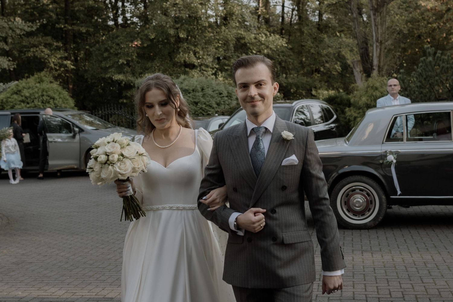A couple walks together at their wedding celebration in a parking area with vintage cars visible in the background.