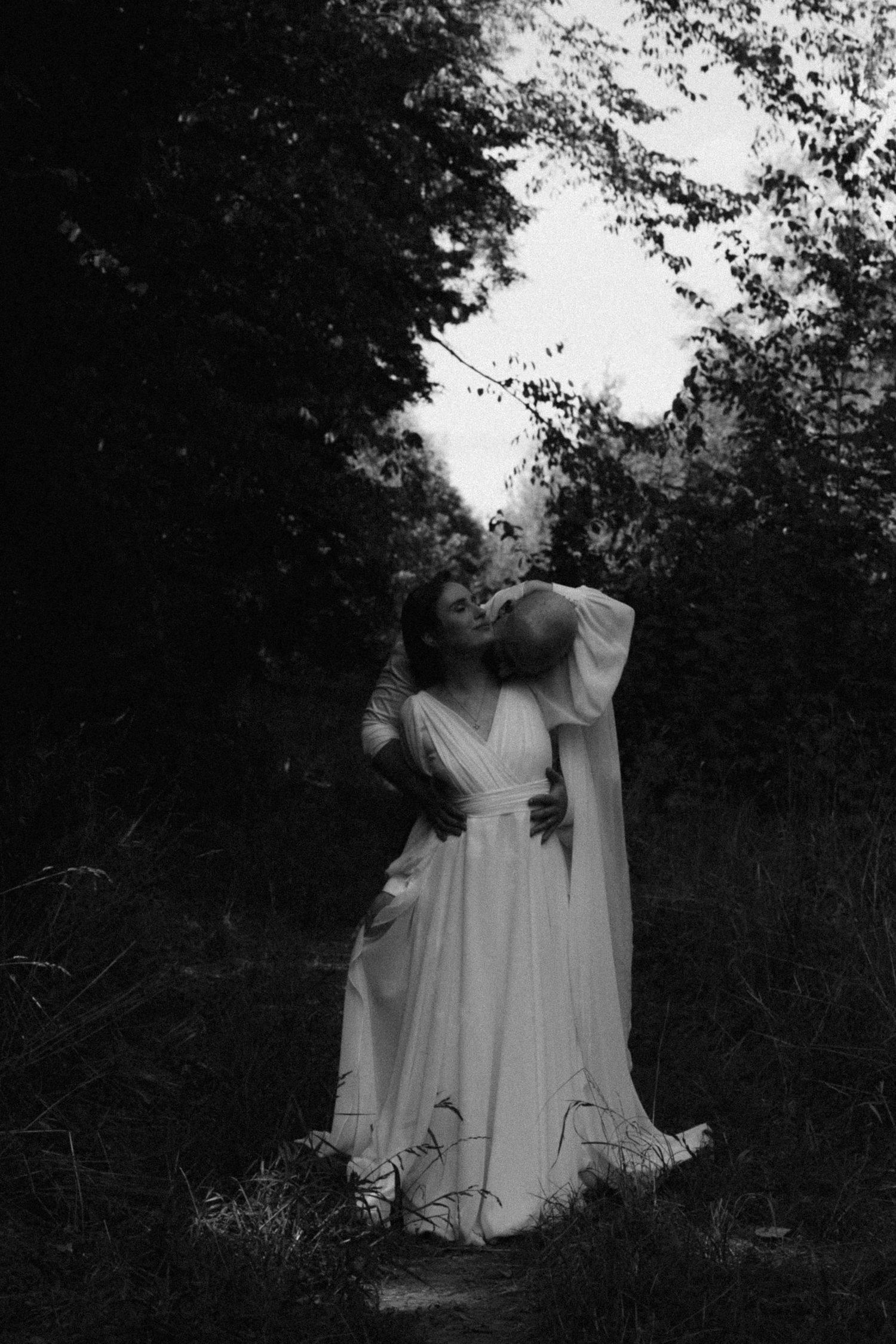 A ghostly figure in white dress stands among dark trees in an atmospheric black and white forest scene.