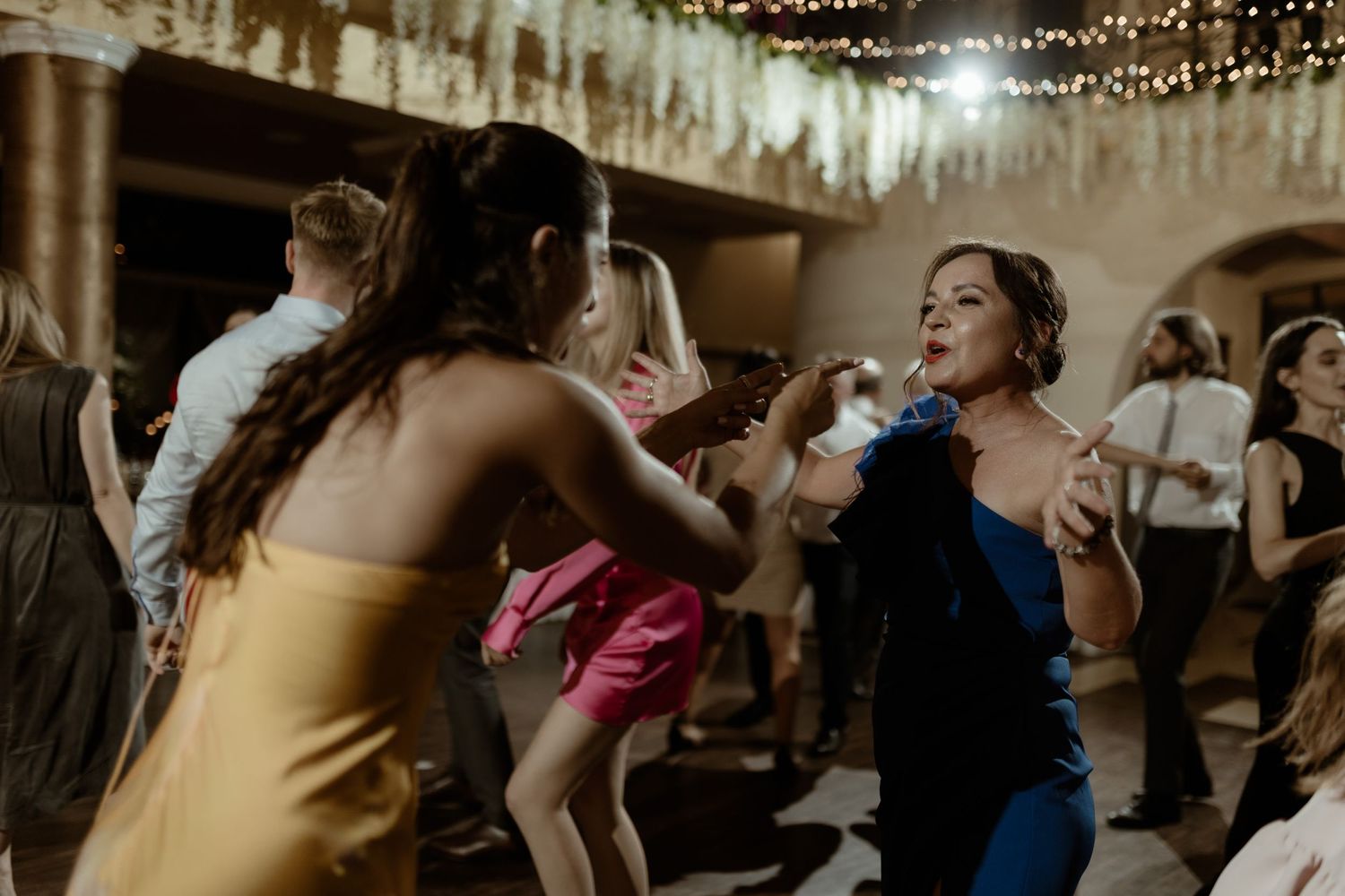 People dancing with joyful energy at a festive reception under glittering string lights and decorations.
