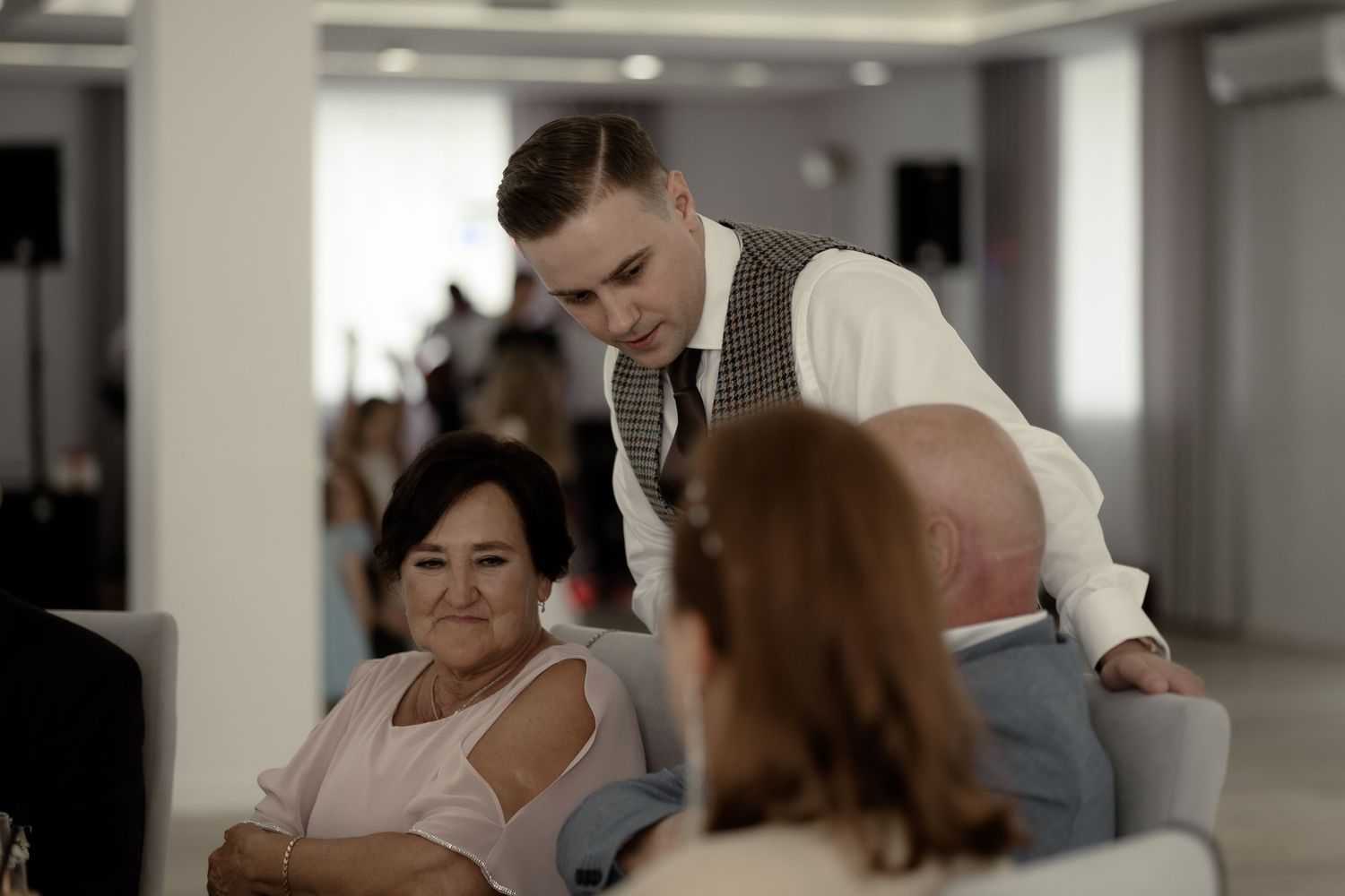 In a bright modern office space, a professionally dressed server in vest and slacks attends to dining guests at their table.