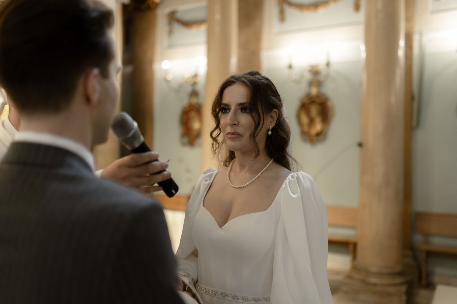 A bride in white dress listens intently during wedding ceremony as groom speaks into microphone in elegant venue.