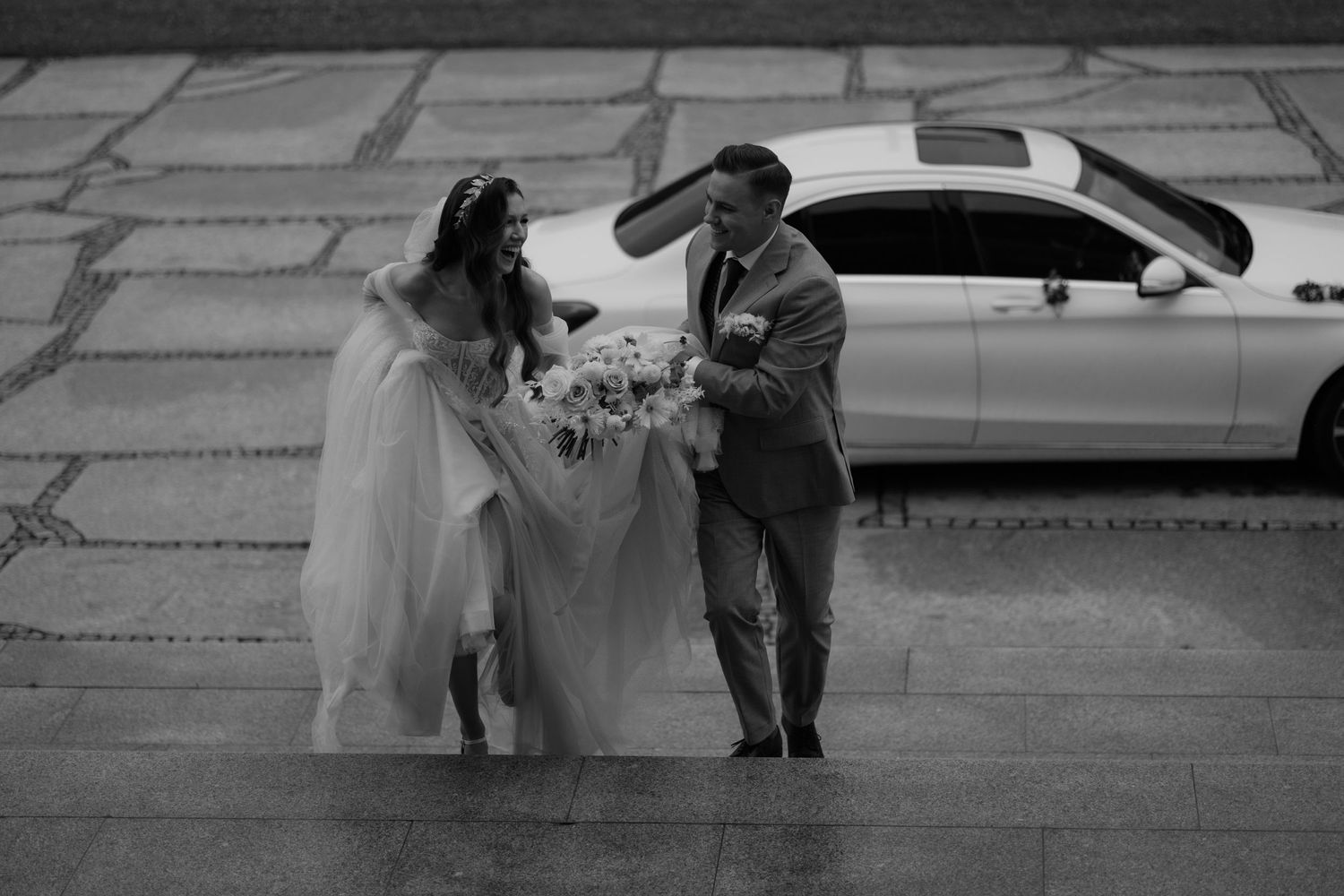 A black and white photograph shows a couple in wedding attire walking together outdoors near a parked car.