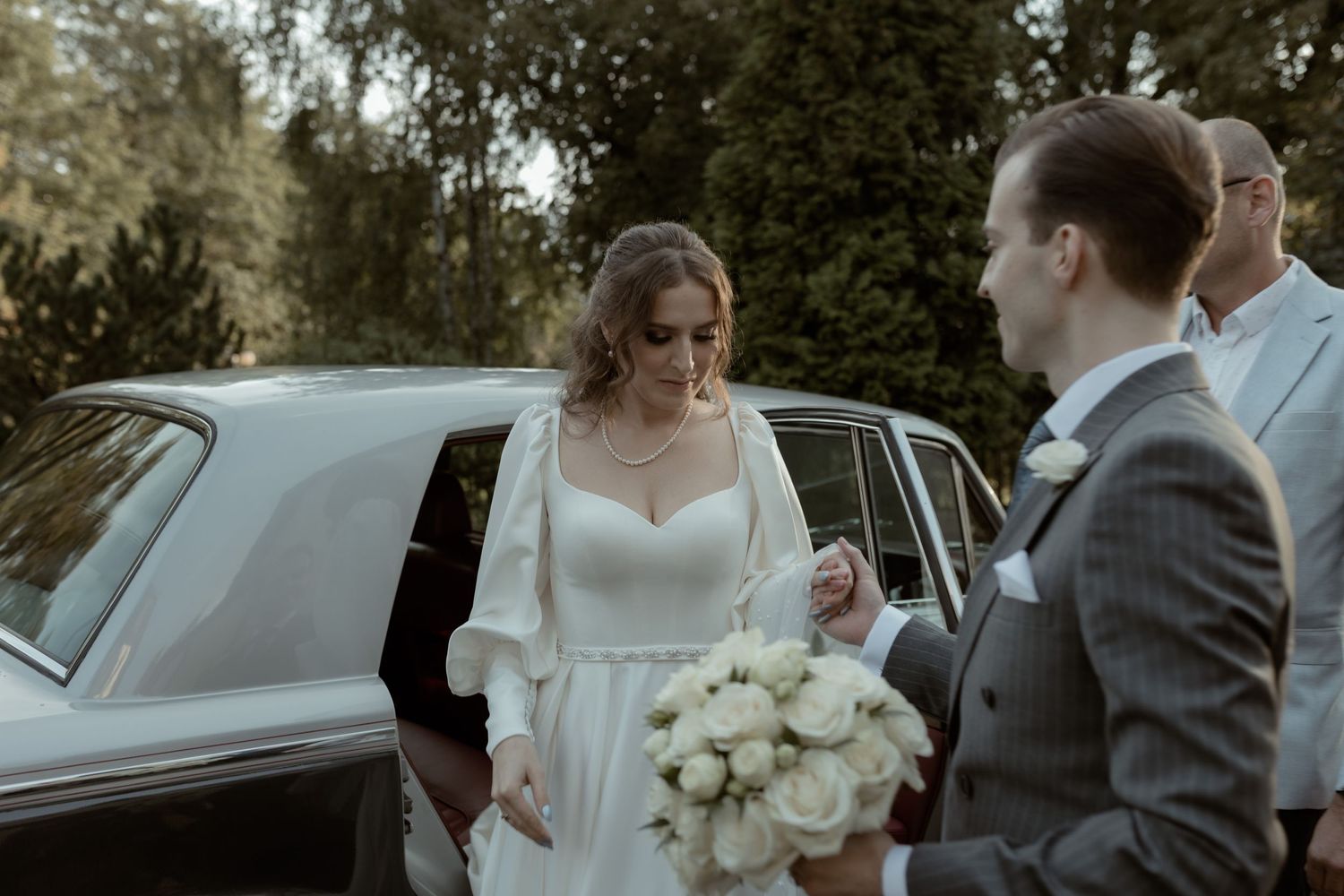 A couple shares an intimate moment during their wedding ceremony next to a classic car while holding white roses.