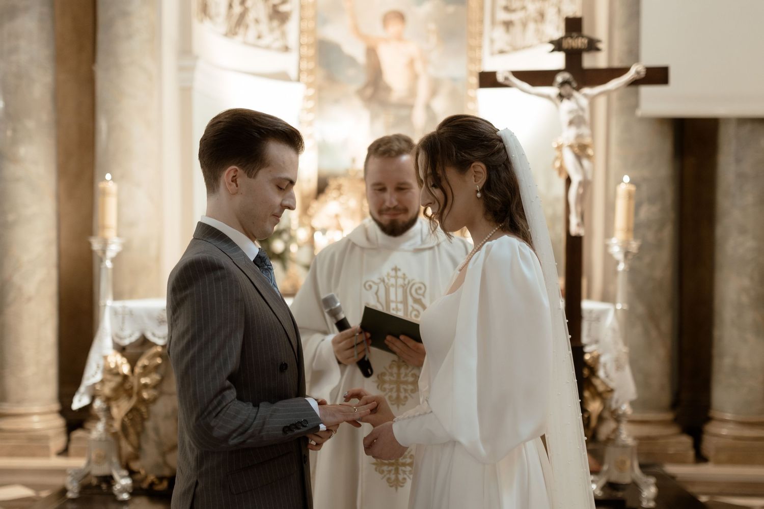 A couple exchanges wedding vows during a traditional church ceremony with ornate decor and a crucifix in the background.